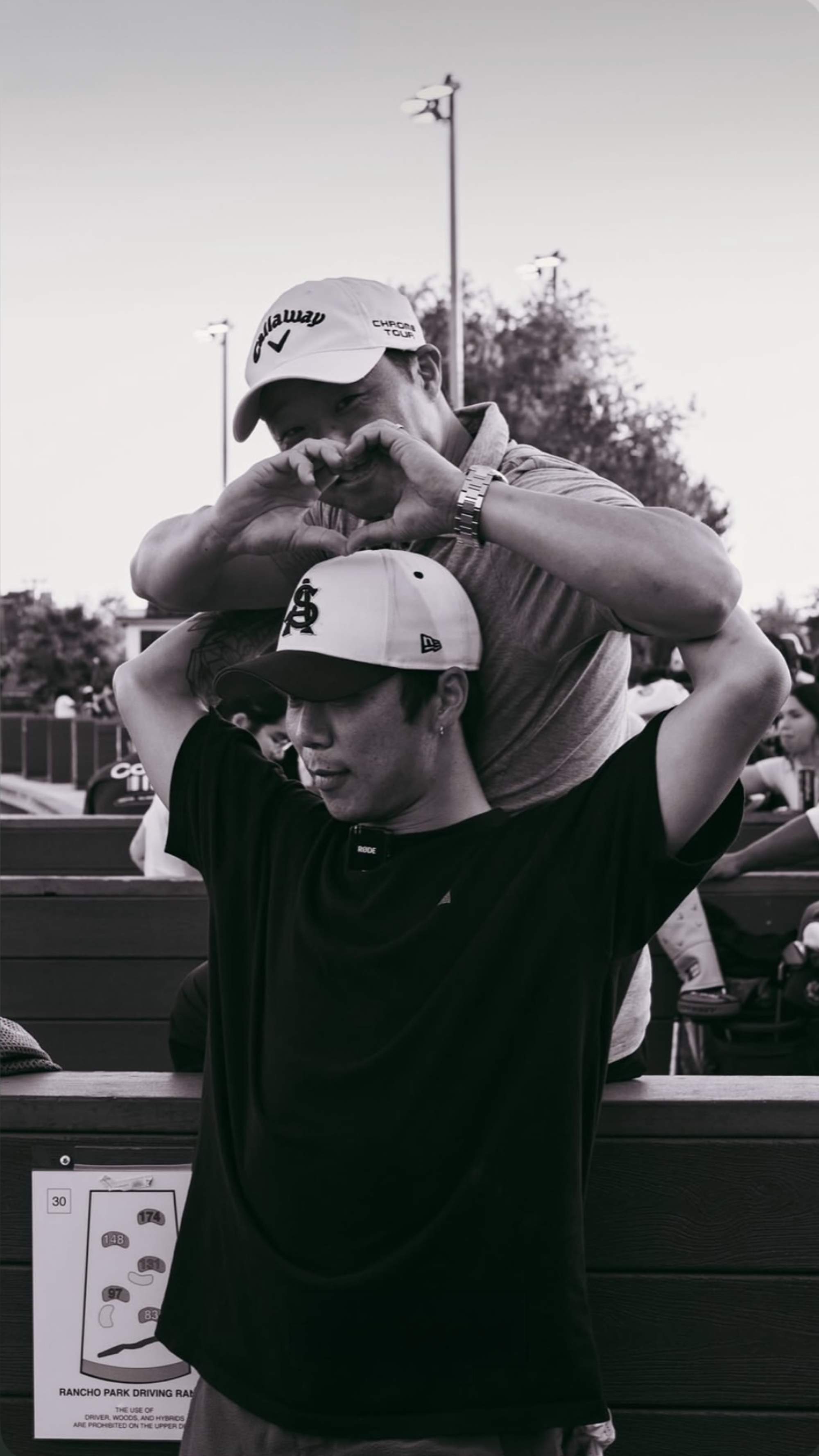 A person wearing a black T-shirt and a cap with the New York Yankees logo is making a heart shape with their hands. Another person, on top of them, wearing a white Callaway cap and a watch, is also making a heart shape with their hands. They are at an outdoor event with other people around, and there is a poster with a map of Rancho Park Driving Range on the wall.
