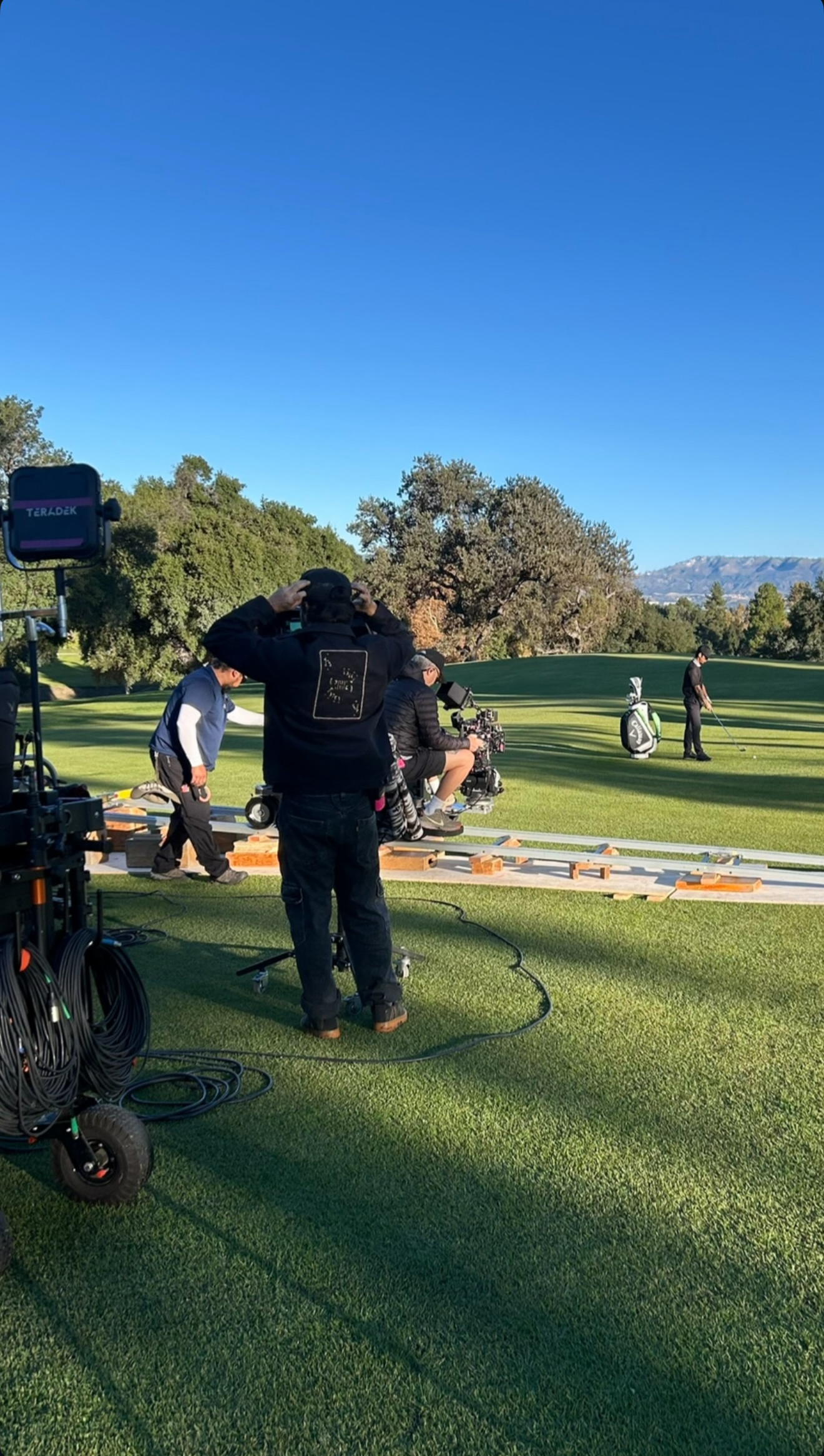 Film crew members on a golf course filming a scene with a man preparing to hit a golf ball, with studio equipment and cameras setup.