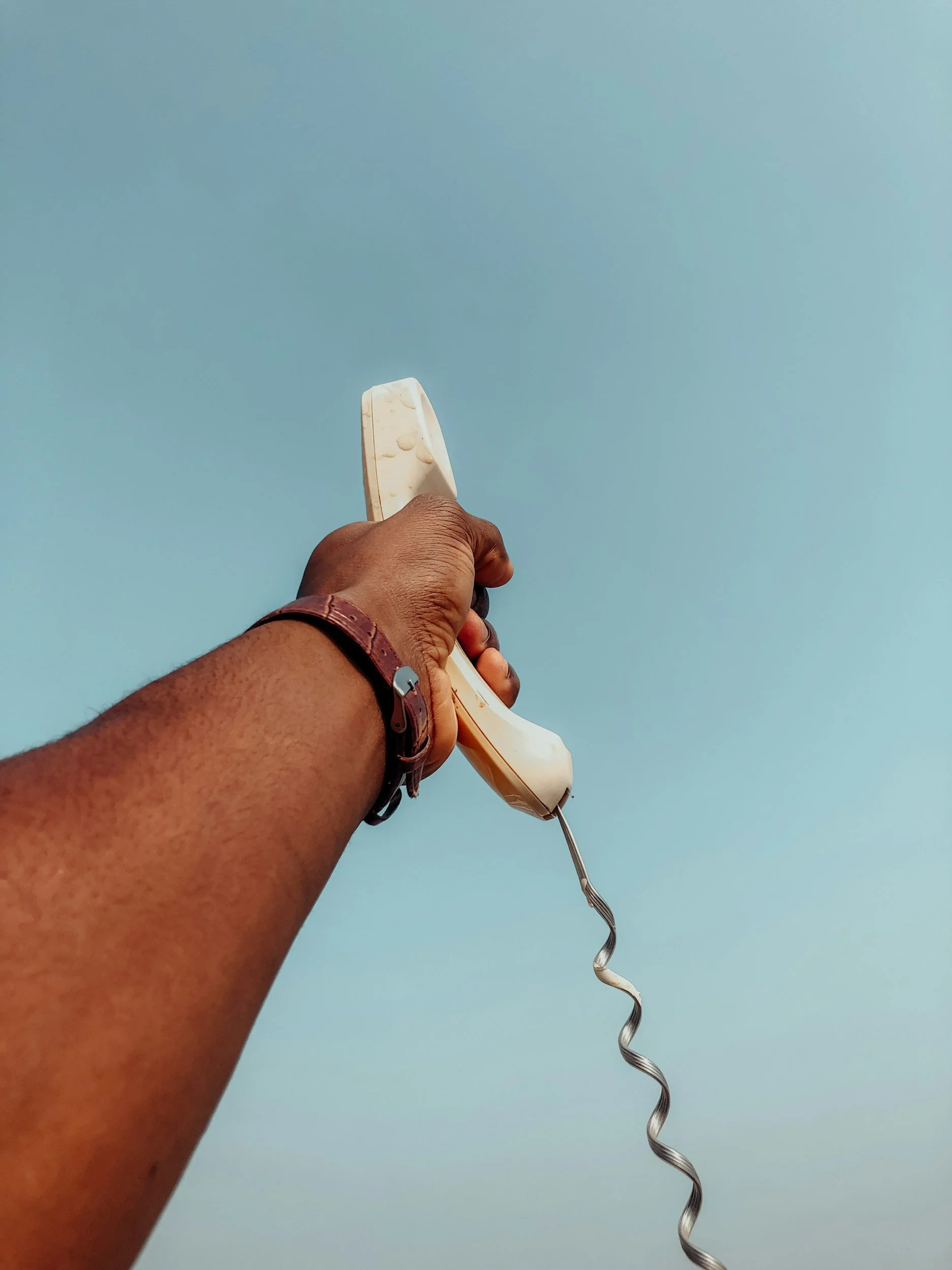 A person's hand holding a traditional white telephone with a spiral cord against a clear blue sky.