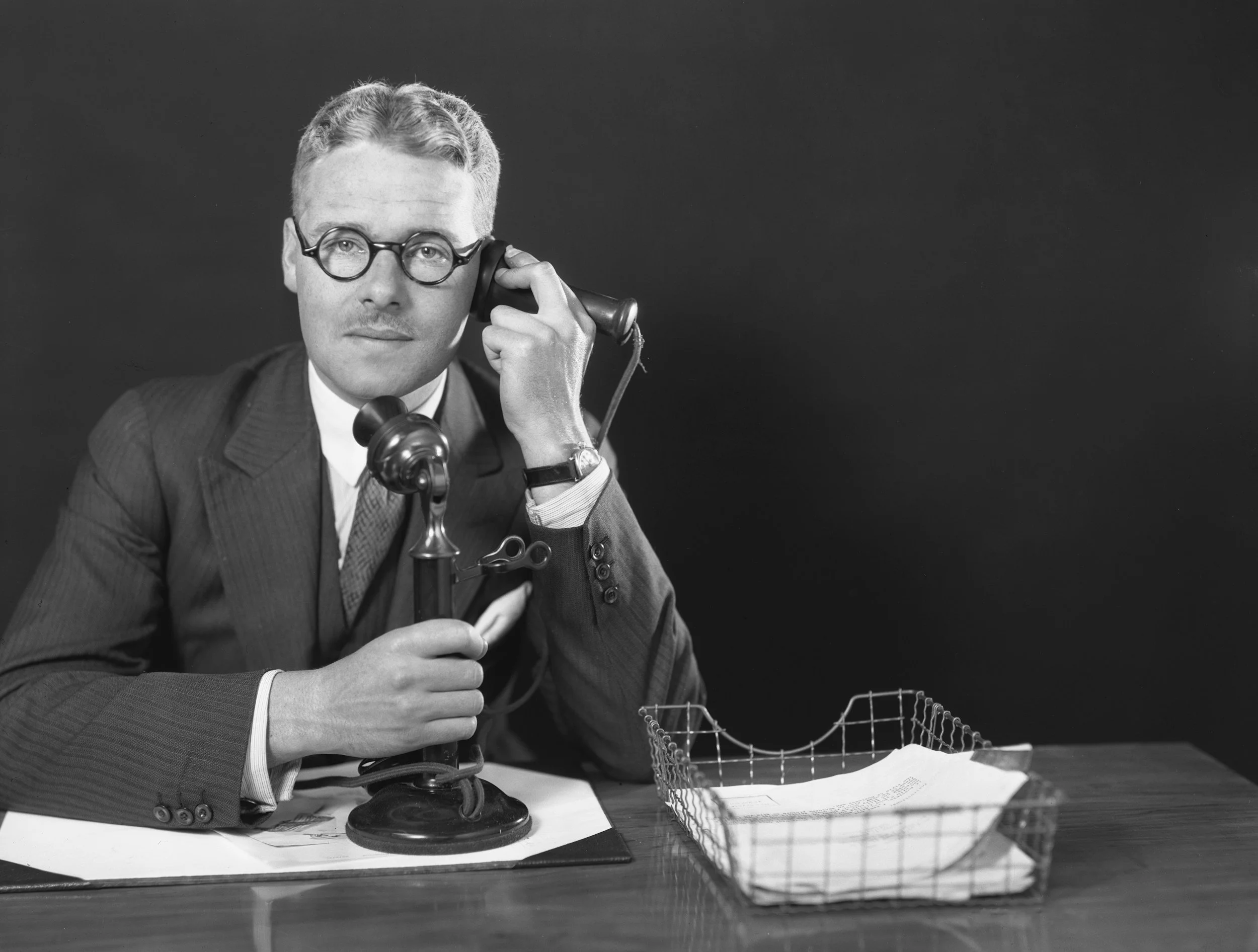 A black and white photo of a man wearing glasses and a suit, holding a telephone receiver to his ear. He is sitting at a desk with papers, and there is a wire rack or tray with documents and files on the desk.