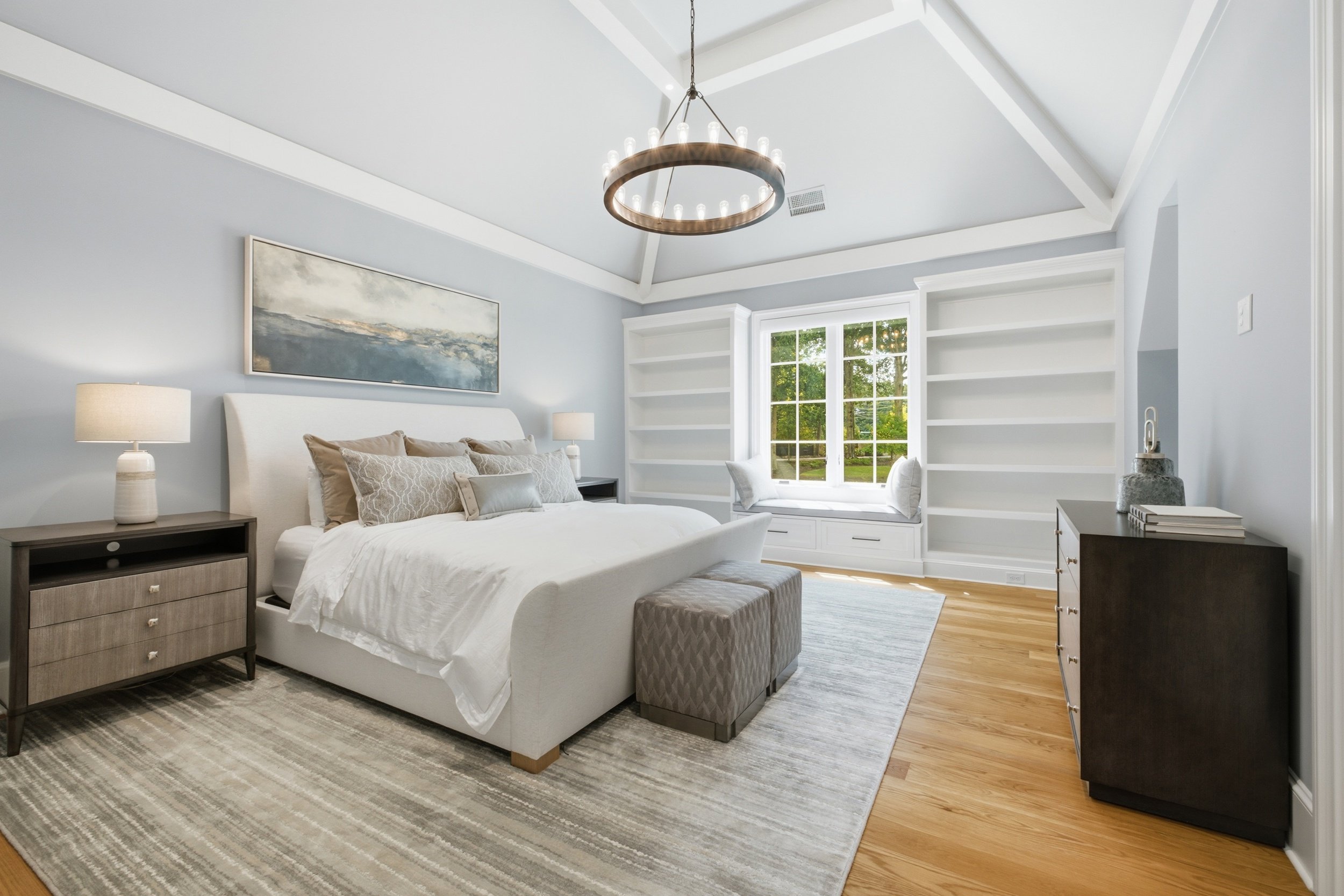 Modern bedroom with white walls, a bed with neutral-colored bedding, two nightstands with lamps, built-in shelves by a window, and a circular chandelier hanging from the ceiling.