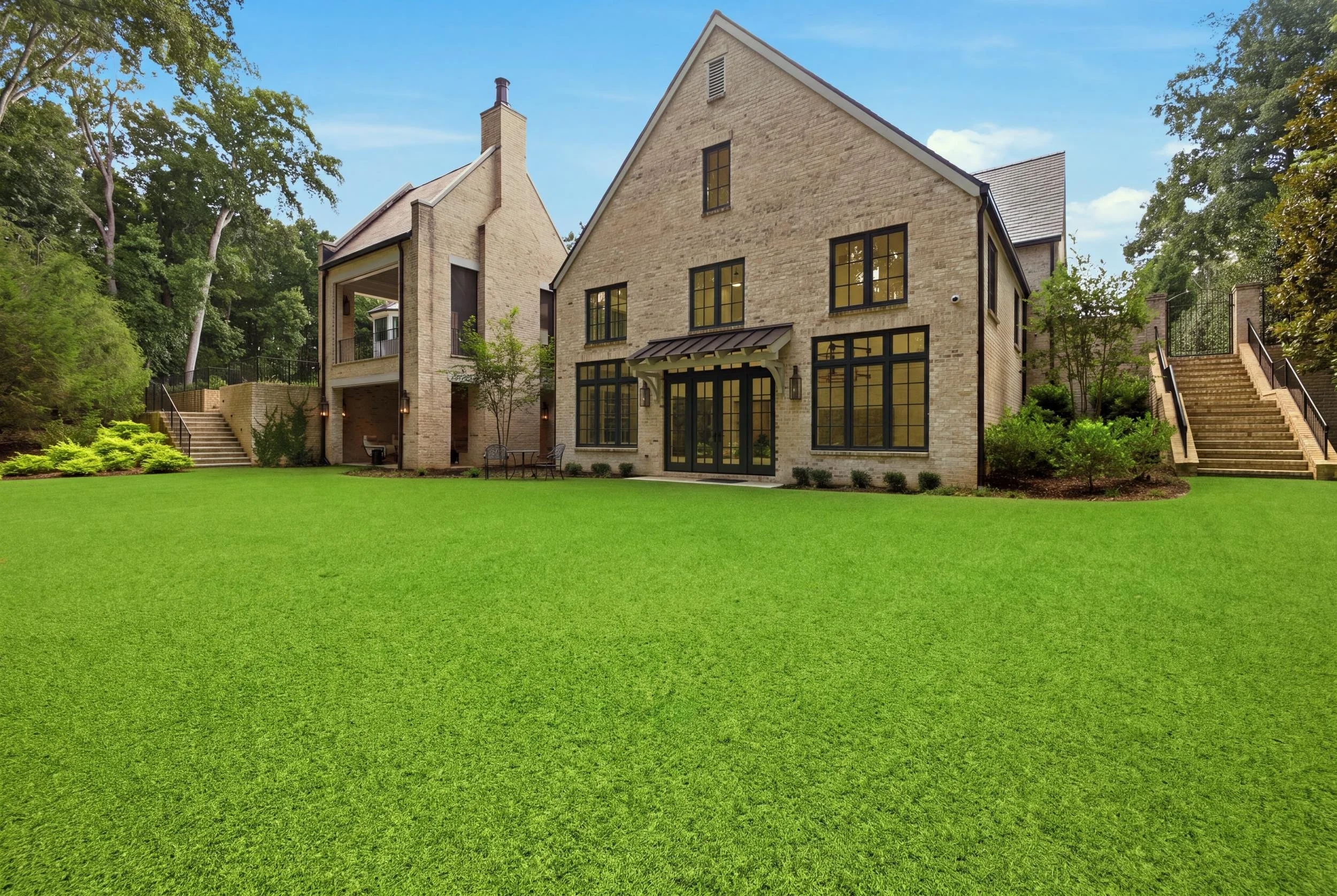 A modern brick house with large black-framed windows and a steep gabled roof, with exterior stairs on either side leading to the backyard, surrounded by green trees and a lush lawn under a partly cloudy sky.