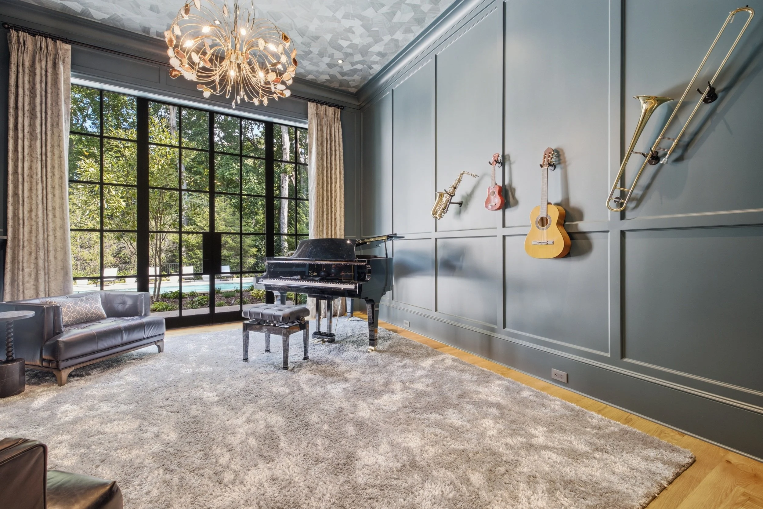 Living room with large window, curtains, chandelier, gray wall with musical instruments including a saxophone, ukulele, guitar, and trombone hanging on it, black piano, vintage grey armchair, and beige carpet.