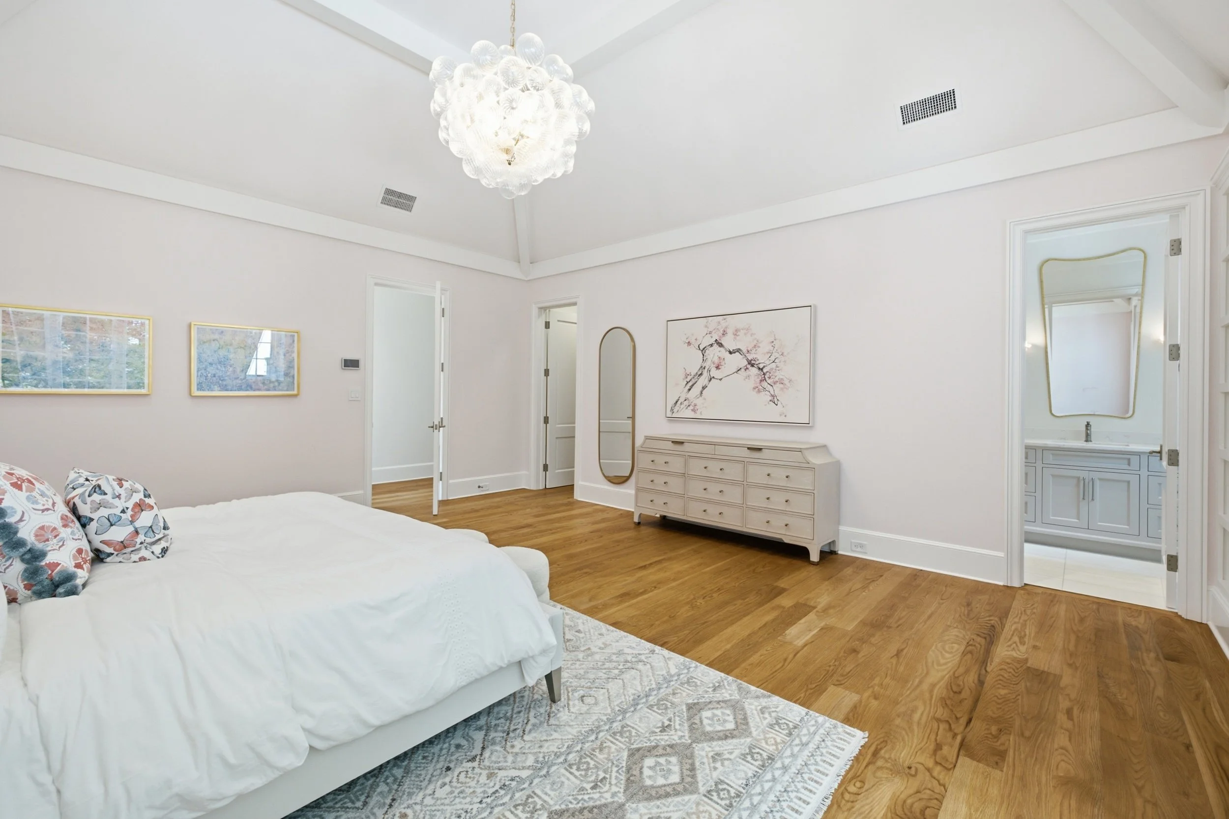 A bright, modern bedroom with white walls, a chandelier, a dresser with a mirror, framed artwork, and a bed with floral pillows.