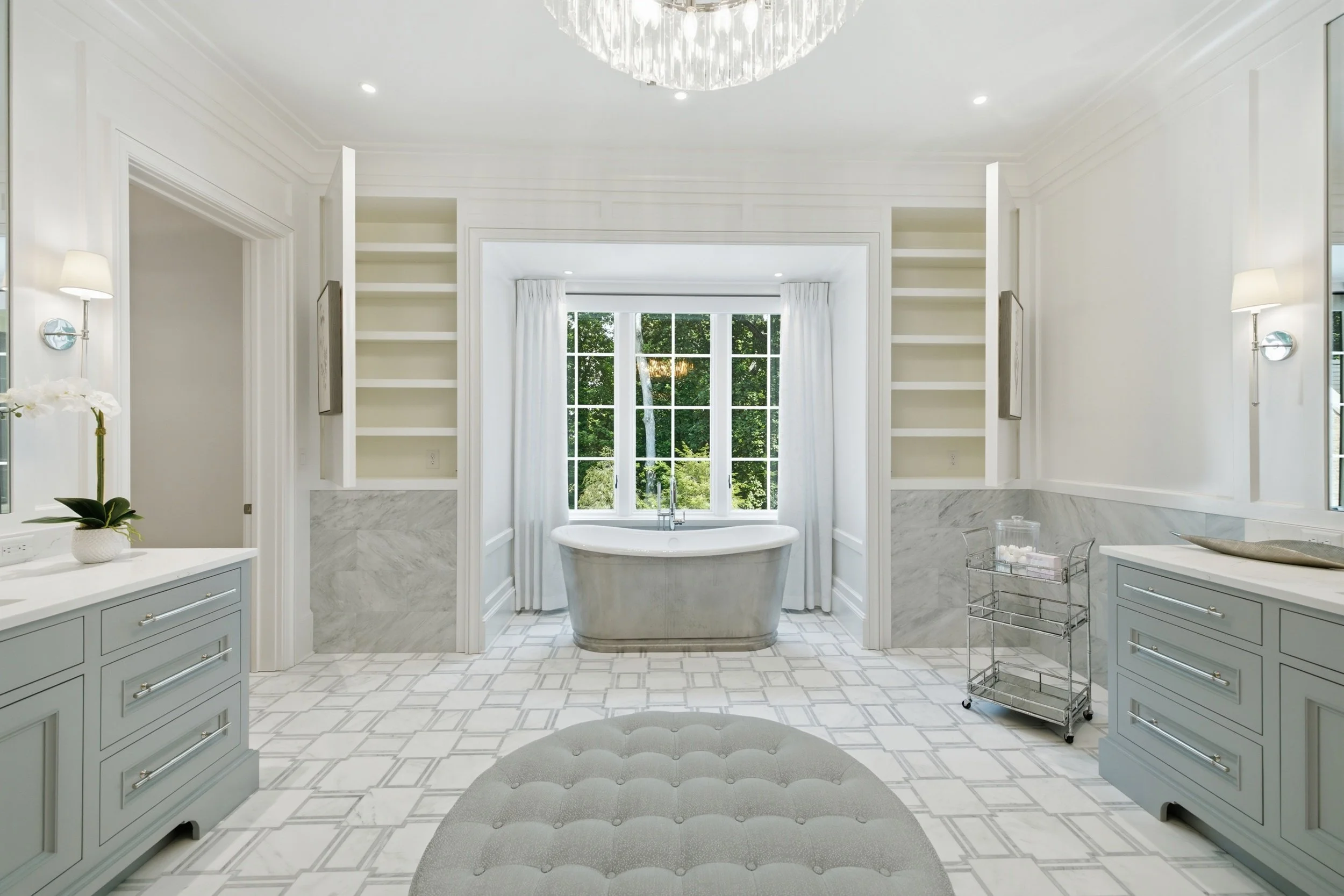 Bright bathroom with a freestanding bathtub beneath a large window, white cabinetry, marble accents, and a chandelier overhead.