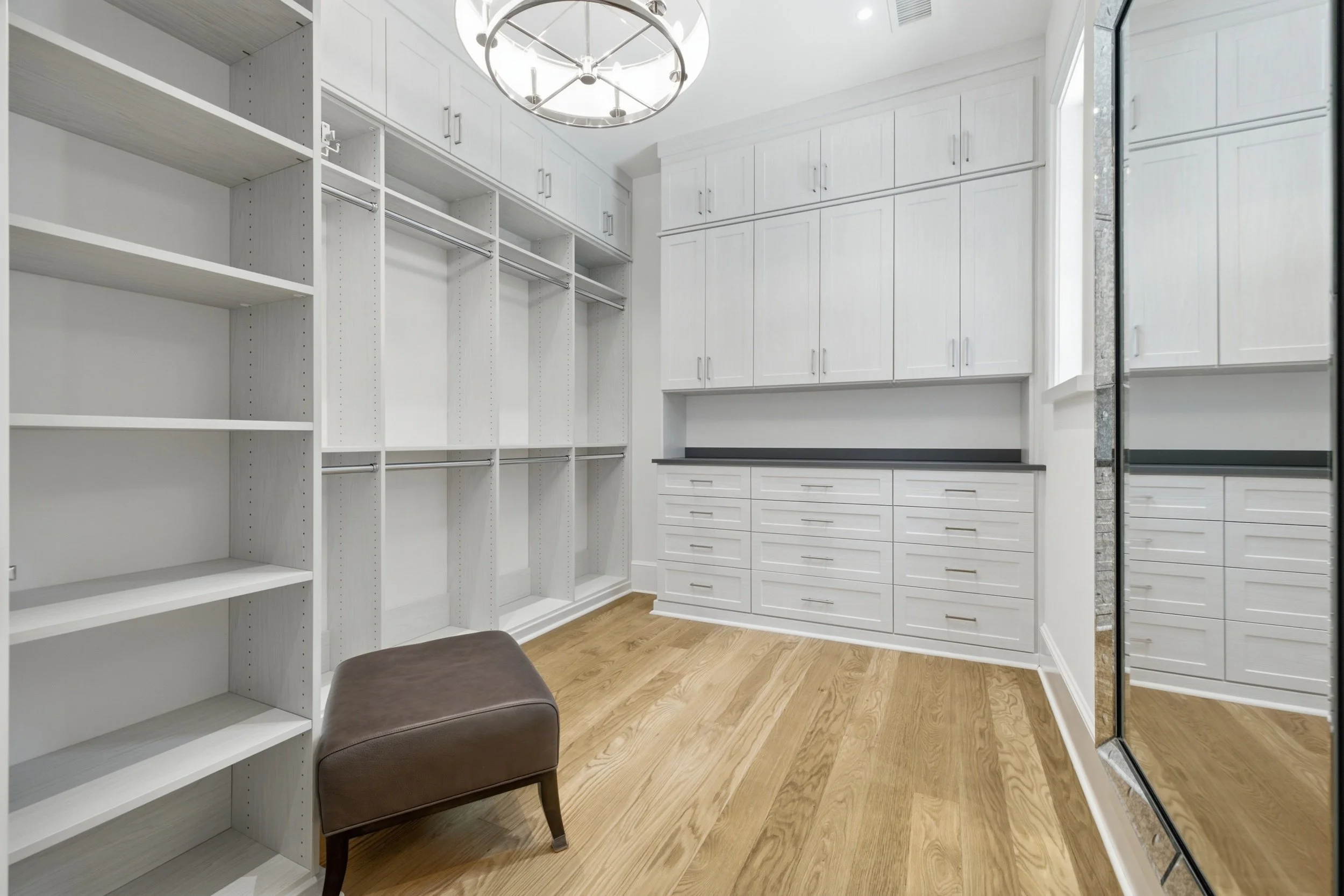 A walk-in closet with white cabinetry, open shelving, multiple drawers, and hanging rods, a brown cushioned stool, wood flooring, and a large window.