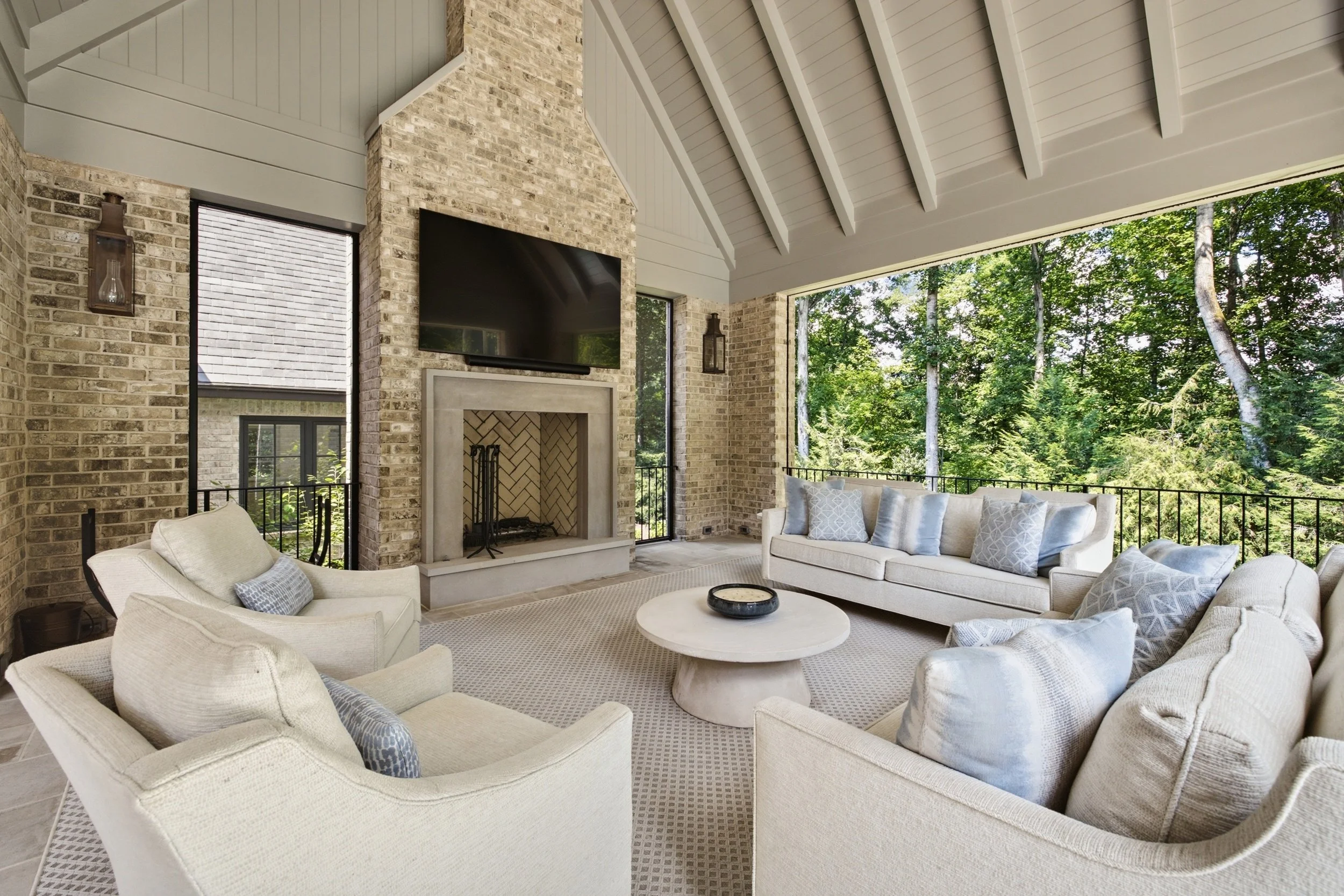 Indoor living space with beige sofas, a round white coffee table, a brick fireplace, a flat-screen TV, large windows showing green trees outside, and a high, white vaulted ceiling.