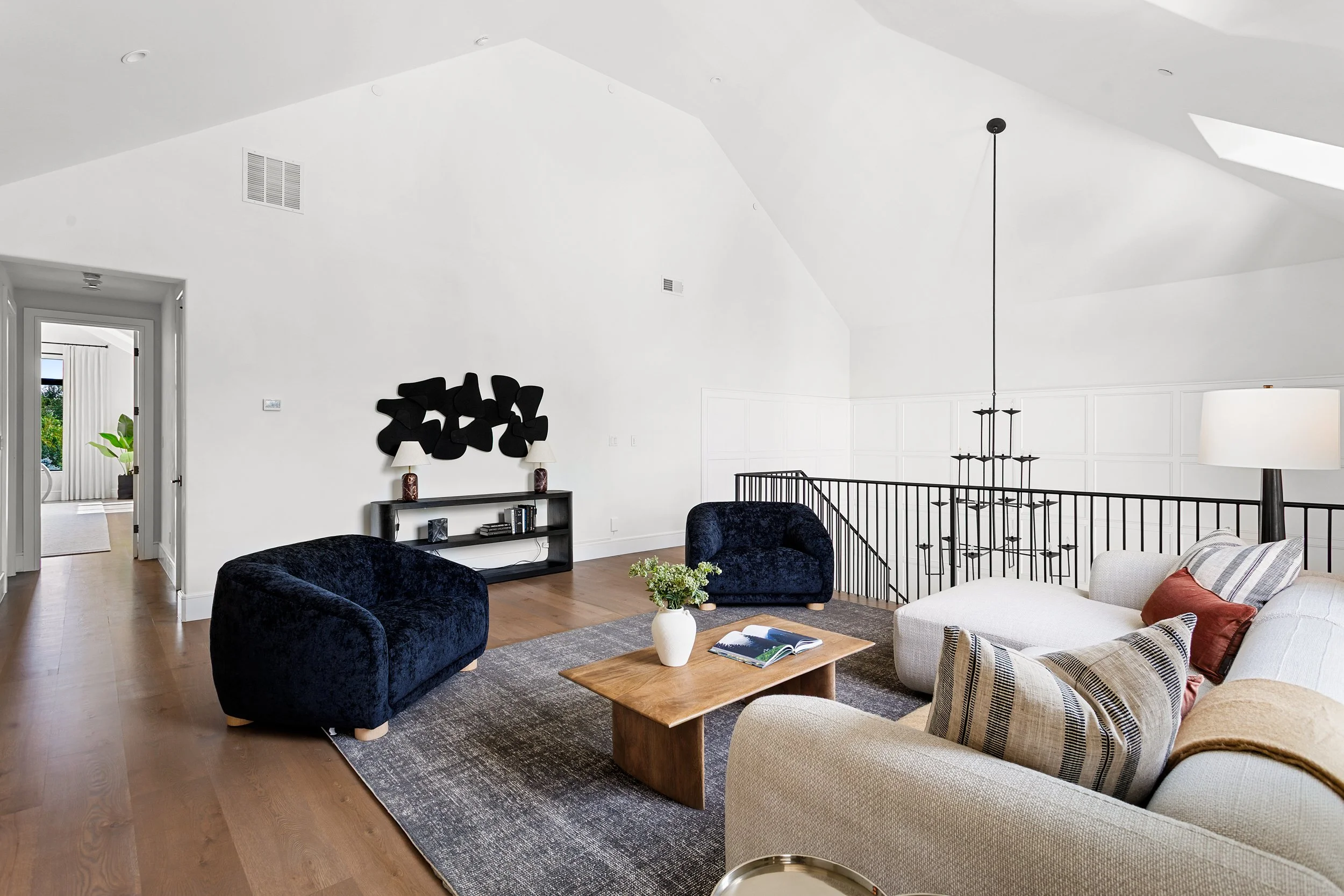A spacious living room with white walls and vaulted ceilings featuring skylights. The room is decorated with black and beige furniture, including velvet armchairs, a sofa with striped pillows, and a wooden coffee table with a vase of flowers. There's