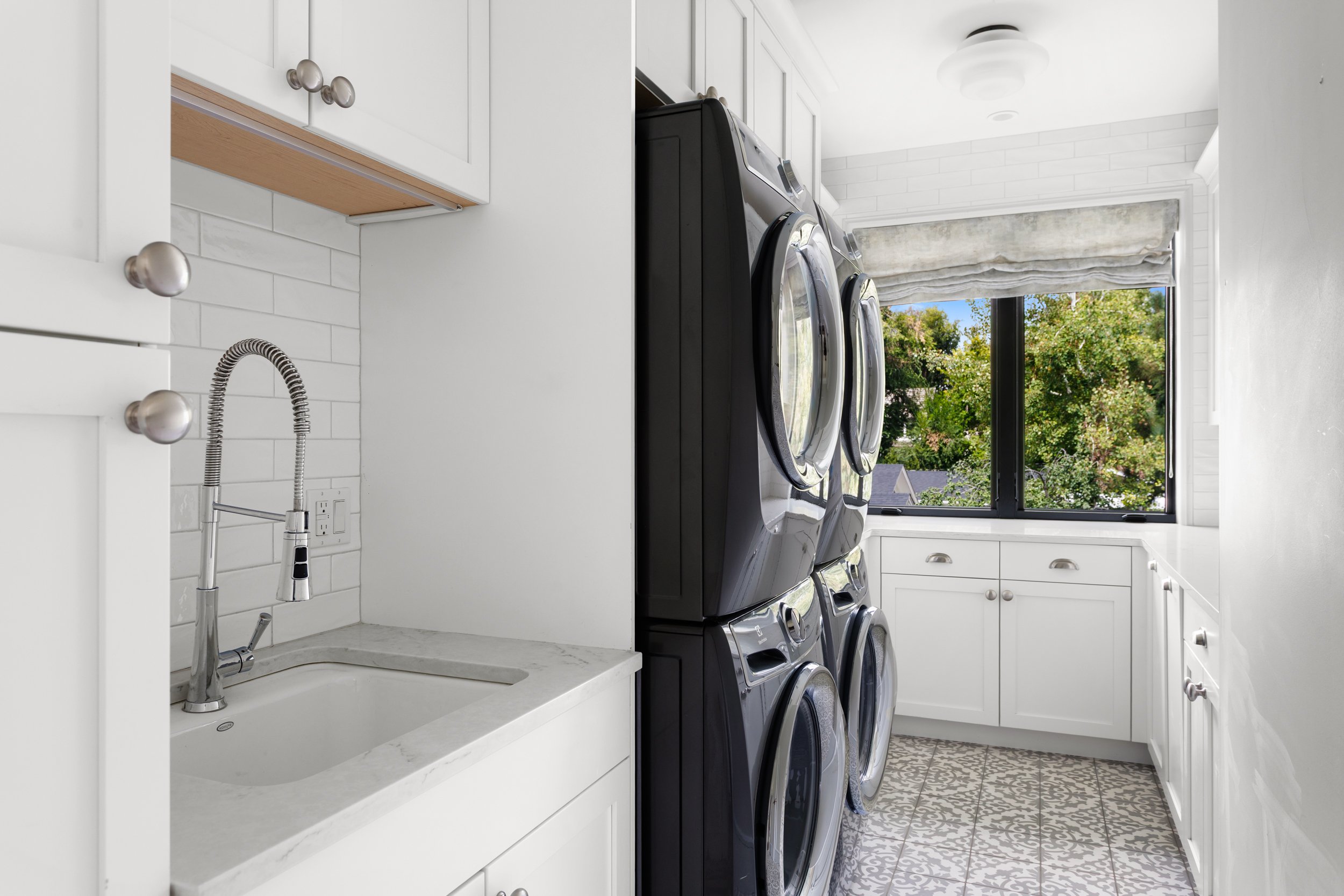 A laundry room with black washer and dryer units, white cabinets, a white marble countertop with a sink, and a window showing trees outside.