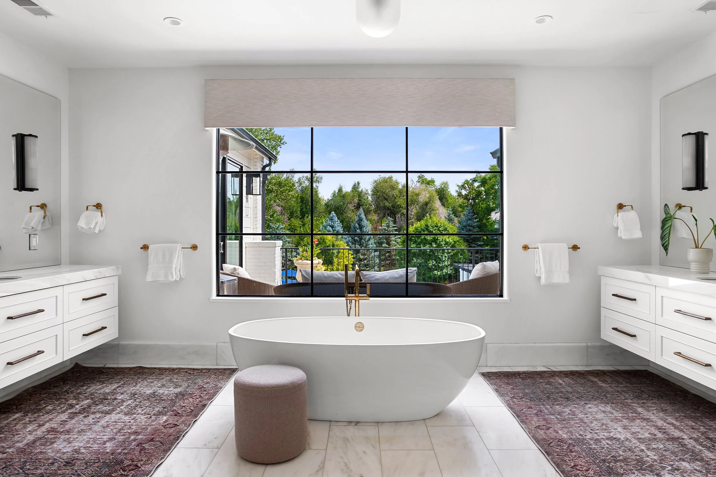 A bathroom with a large window overlooking a green outdoor area, a white freestanding bathtub, two vanities with white cabinetry, and decorative rugs on the marble floor.
