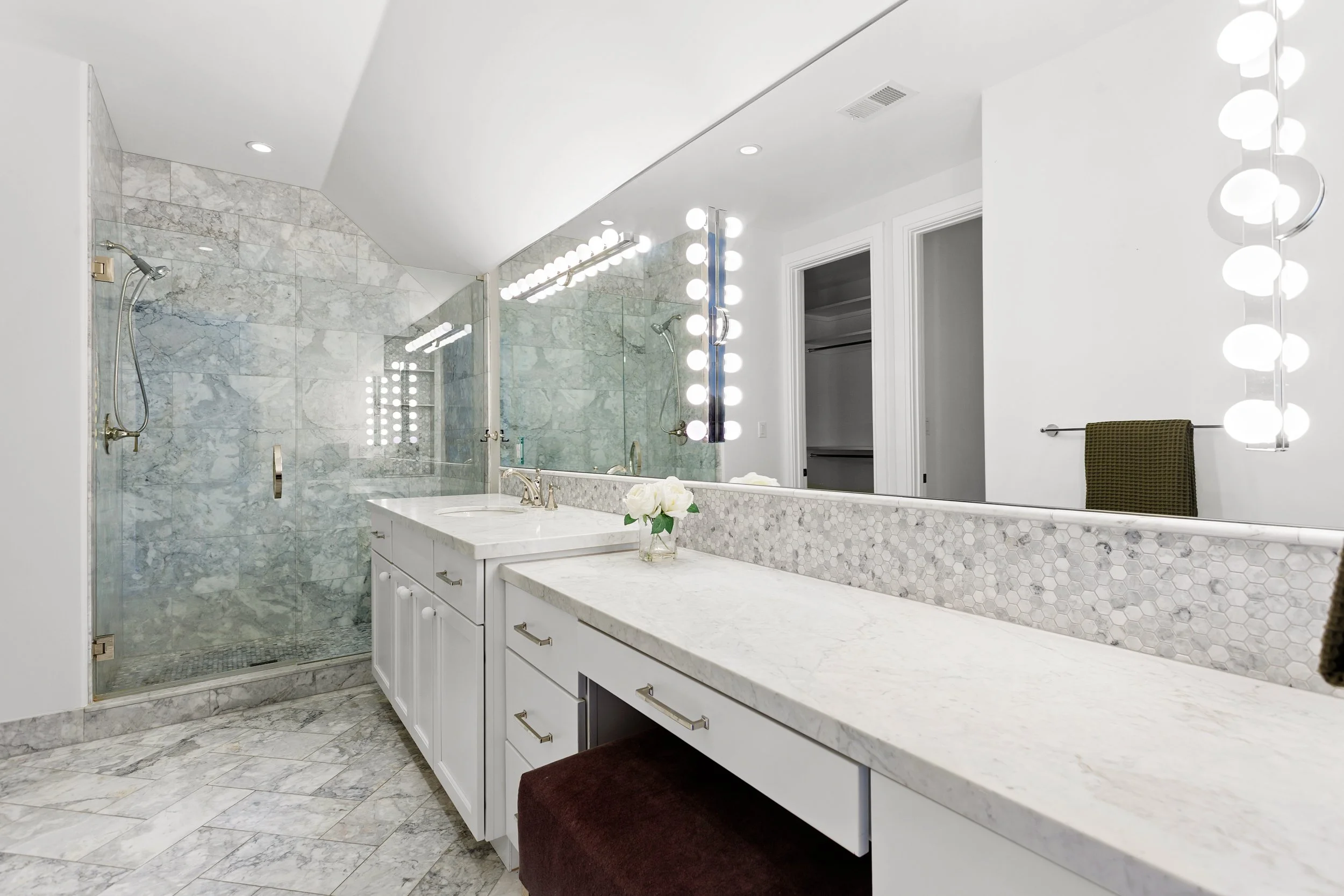 A modern bathroom with a large mirror surrounded by bright vanity lights, a marble countertop with a sink, a glass-enclosed shower with marble tiles, and a towel hanging on a rack.