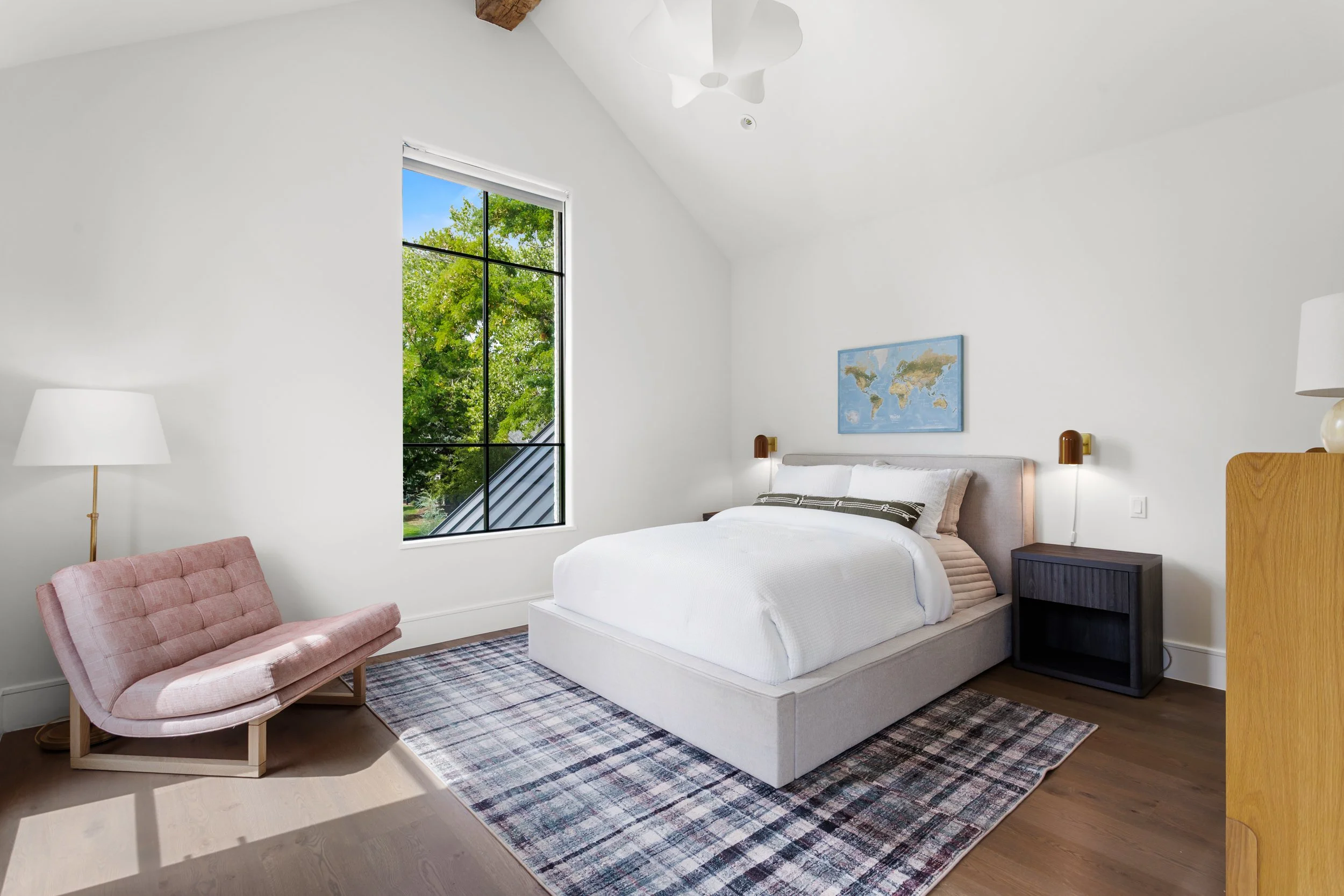 A bedroom with a white bed, a pink upholstered lounge chair, a world map on the wall, large window showing green trees outside, a table lamp, and a plaid rug.