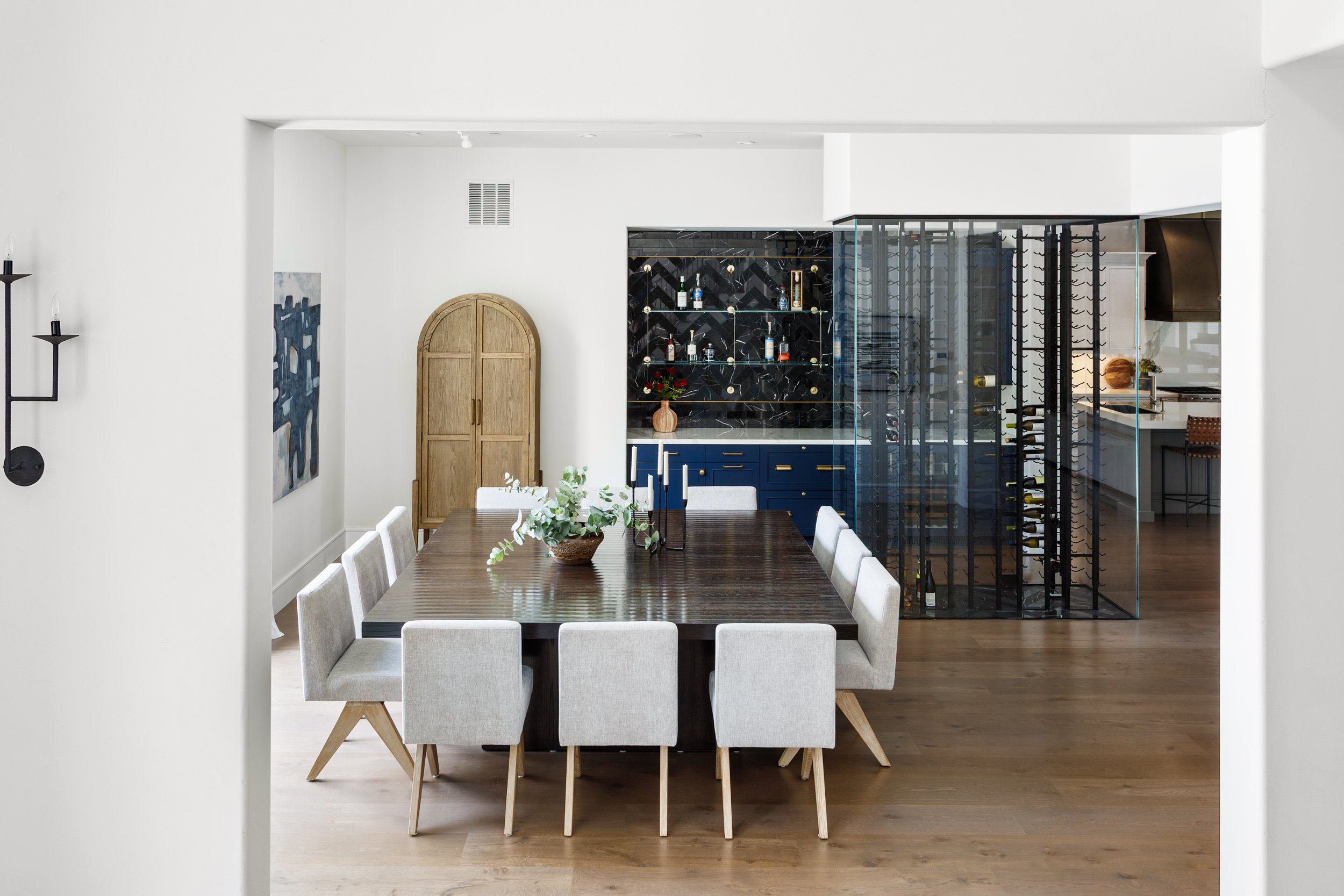 A dining room with a wooden table, surrounded by eight light-colored chairs, and a decorative centerpiece of flowers. In the background, there is a black and gold bar cabinet with bottles and glasses, a wooden arched door, art on the left wall, and a