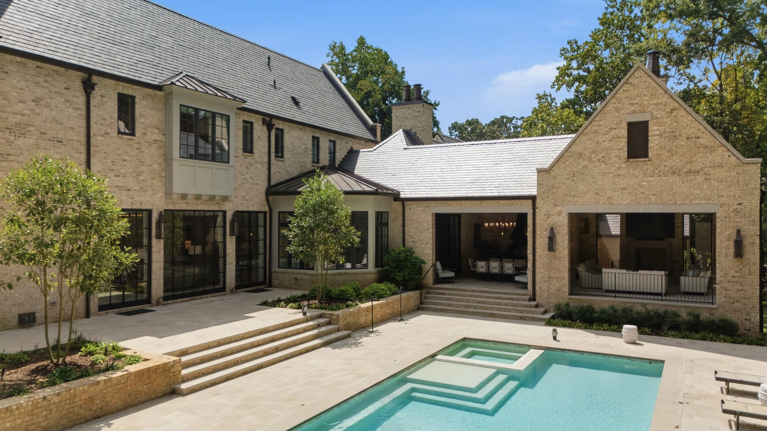 Modern brick house with multiple gables and large windows, backyard with steps, trees, and a pool.