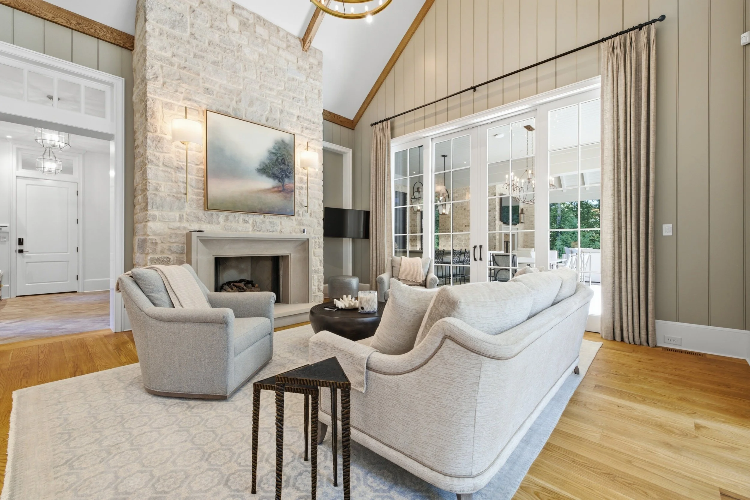 Living room with a beige sofa and armchair, a black circular coffee table with decor, a fireplace with a painting above it, large windows with curtains, and natural wood flooring.