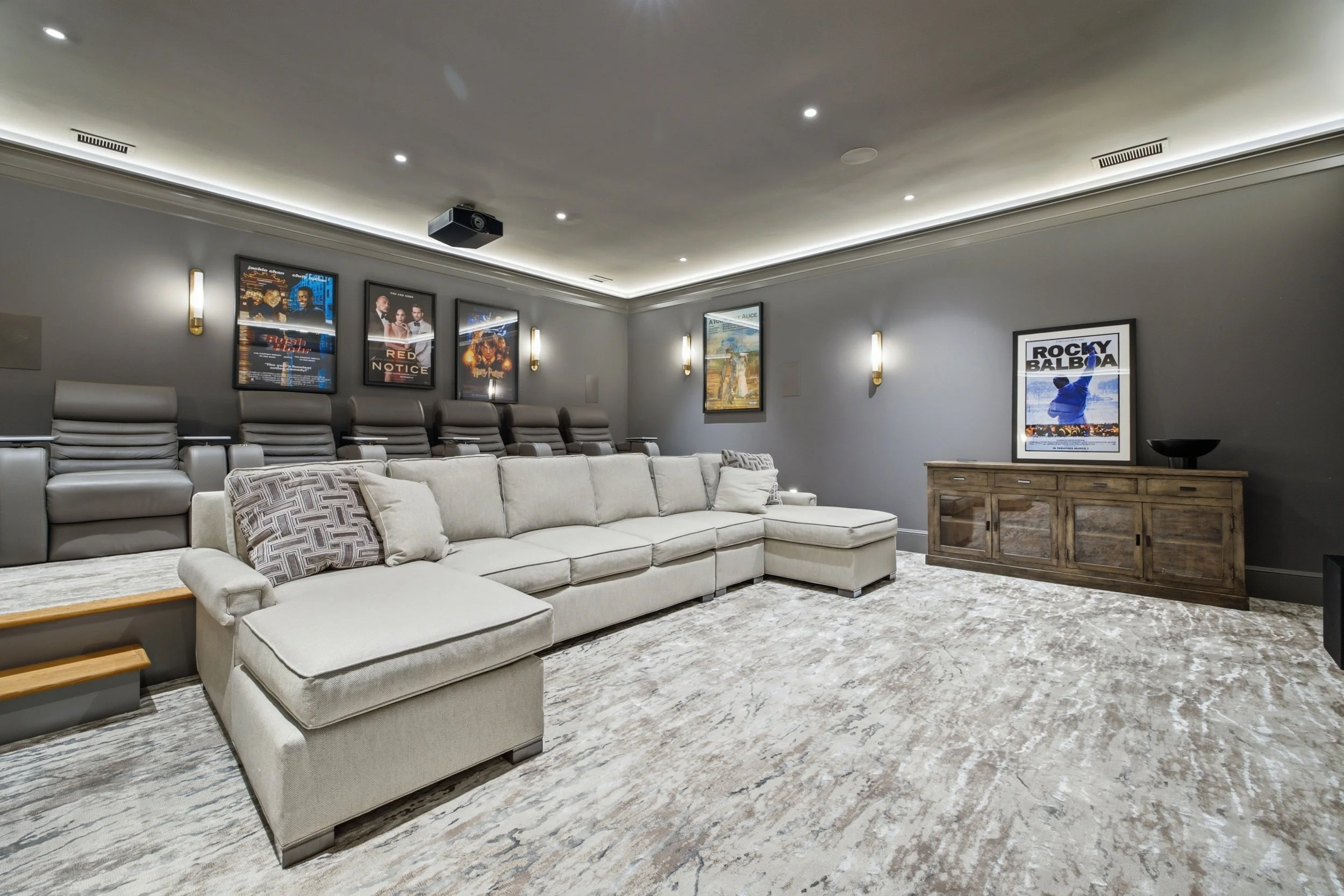 A home theater room with a large beige sectional sofa, gray lounge chairs along the wall, framed movies and posters on the walls, a wooden sideboard, and ceiling lights.