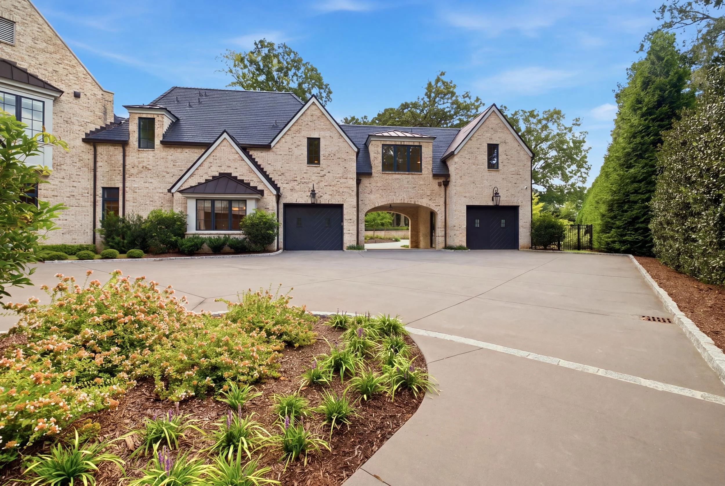 A large, two-story brick house with a black roof, bay windows, and two garage doors, surrounded by landscaped bushes and flowering plants, with tall trees in the background and a peaceful, clear blue sky.