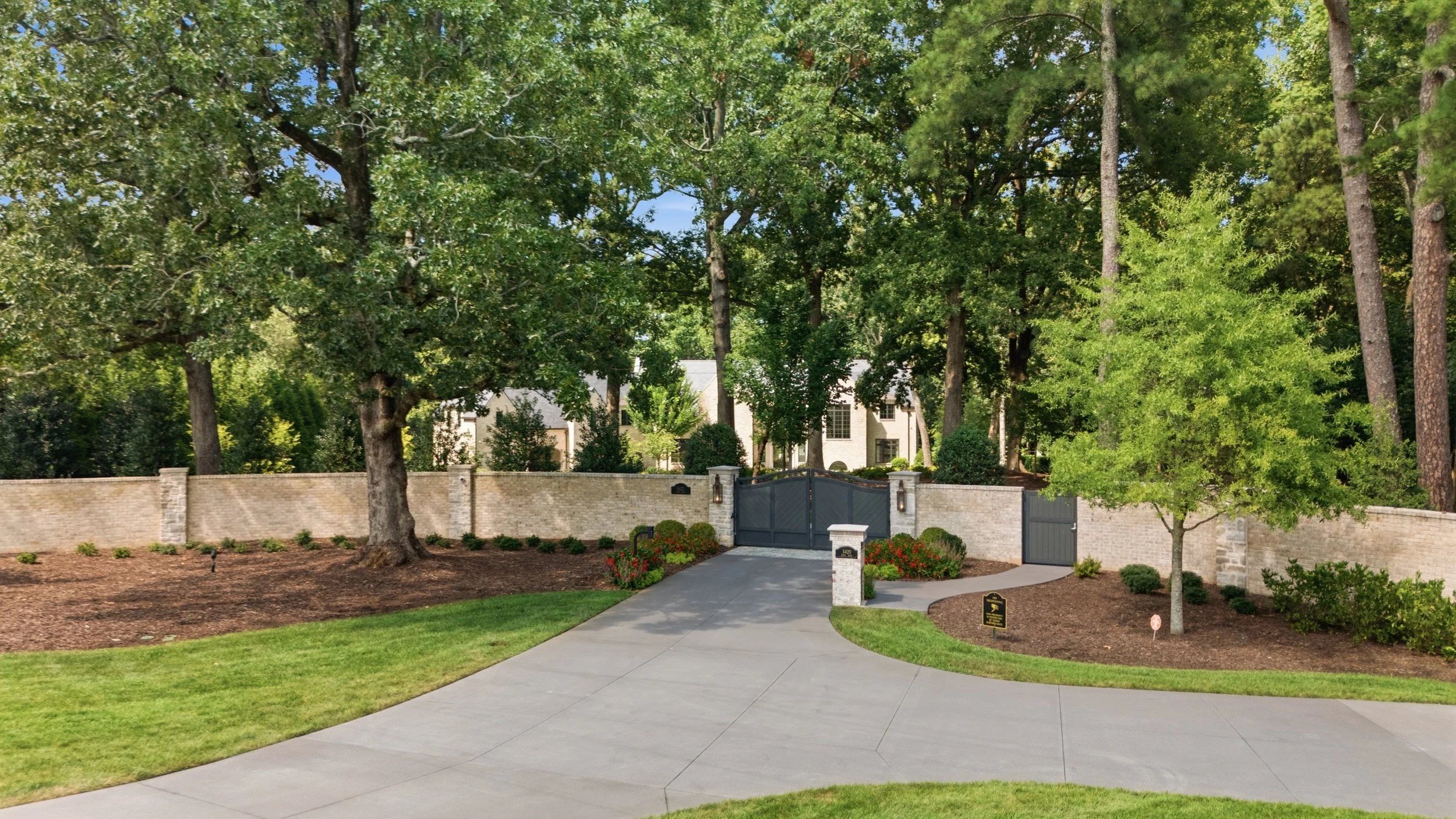 Residential house behind a tall beige brick wall with a black gate, surrounded by trees and landscaped yard with mulch and shrubbery.