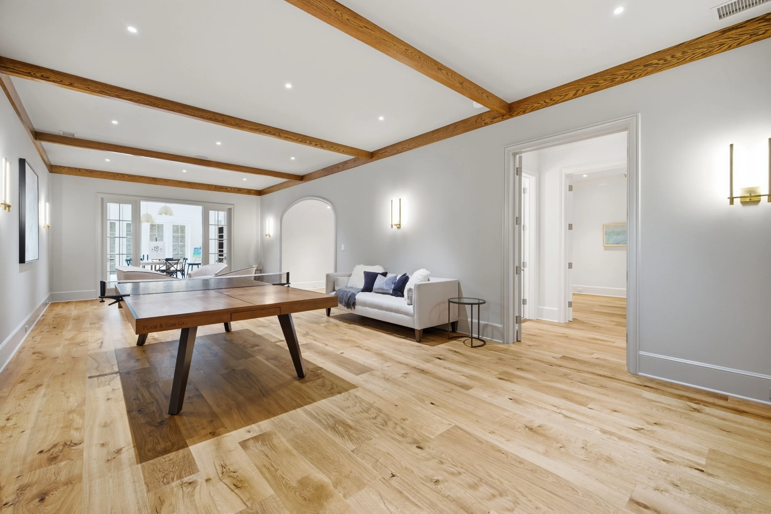 A spacious living area with wooden floors, white walls, and a ceiling with exposed wooden beams. There are recessed ceiling lights, a white sofa with pillows, a small side table, and a dining area with outside access through glass doors.