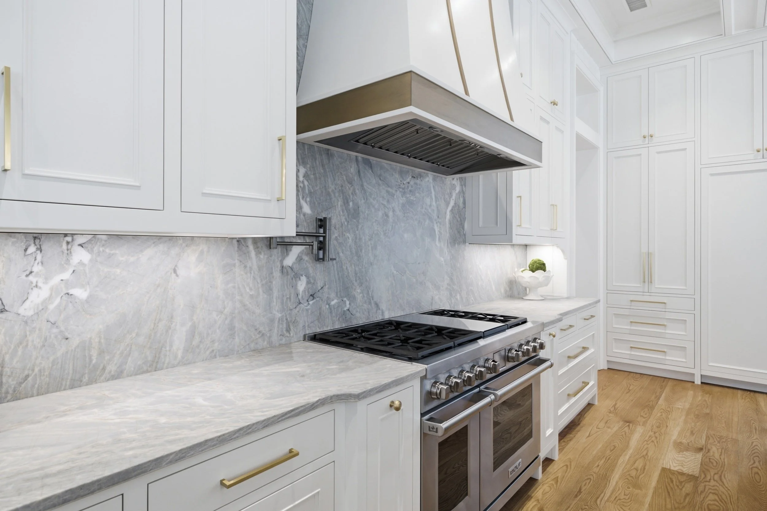 Modern kitchen with white cabinets, marbled gray countertops, stainless steel oven, and gold handles. The kitchen features wooden floors and a white decorative vase with green plants.