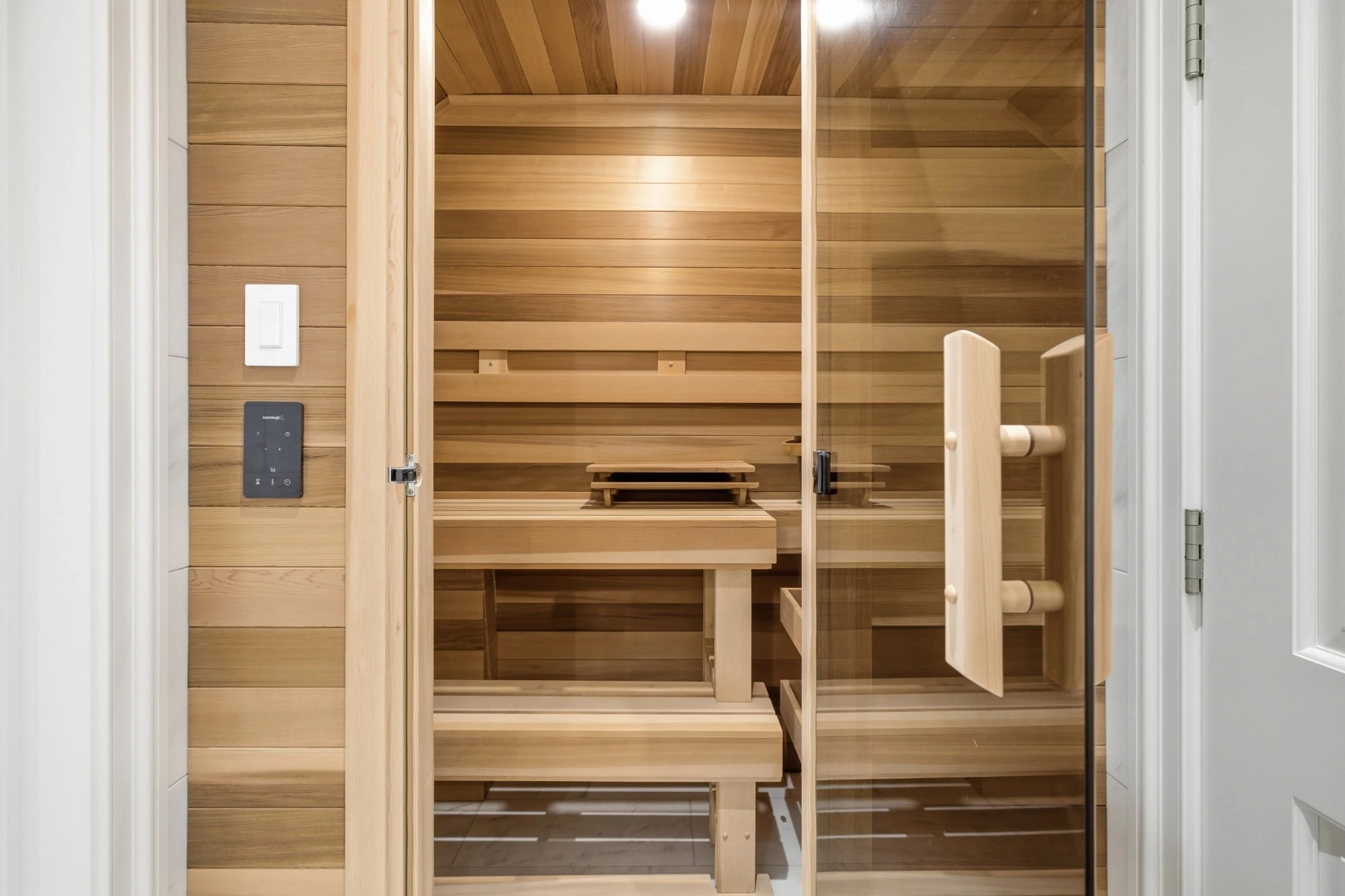 Empty wooden sauna with glass door, wooden benches, and heating controls on the wall.