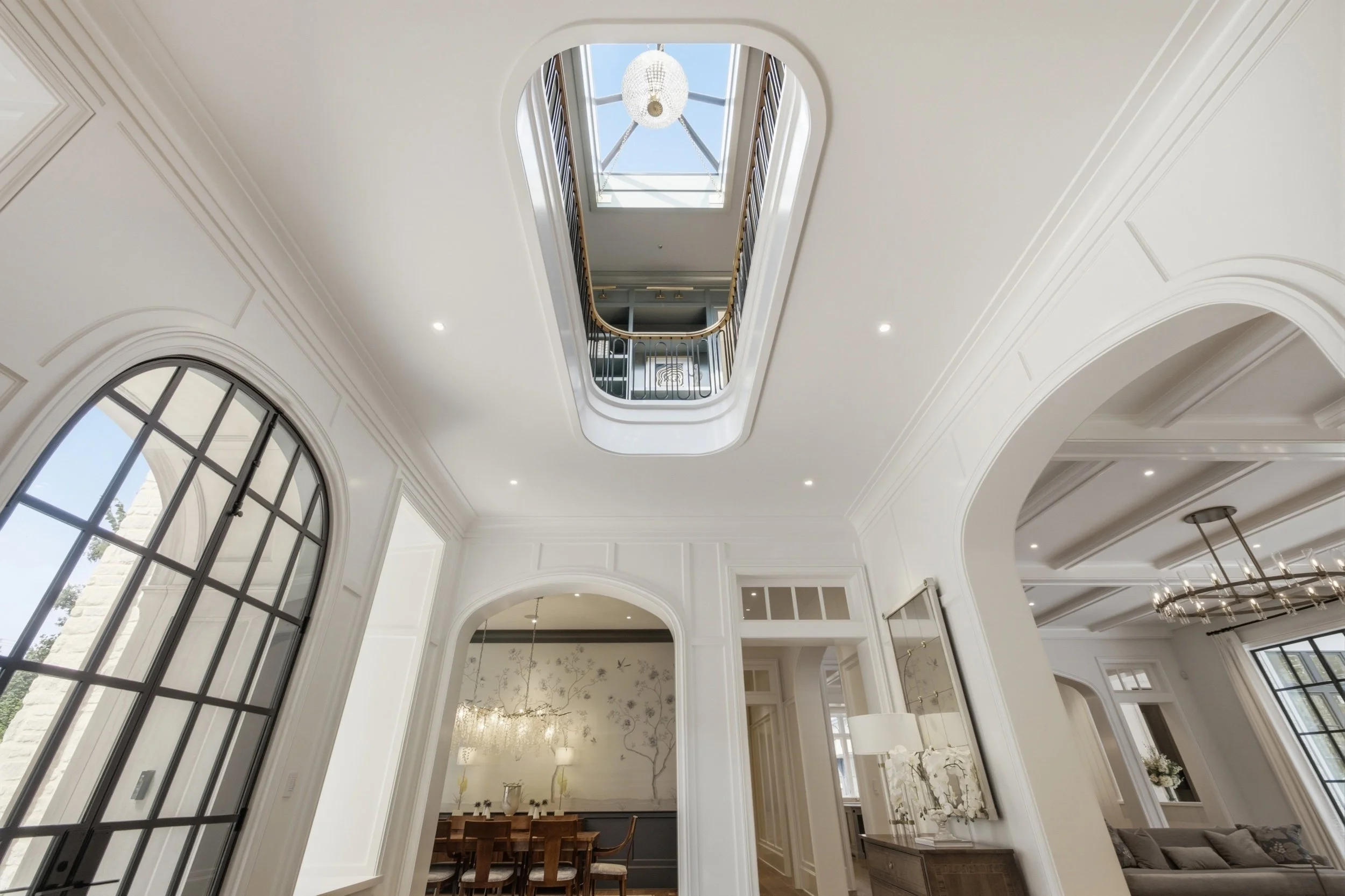 Interior view of a multi-story house with white walls, large arched windows, and a staircase with a black railing leading up to a skylight. The ceiling has recessed lighting and a chandelier.