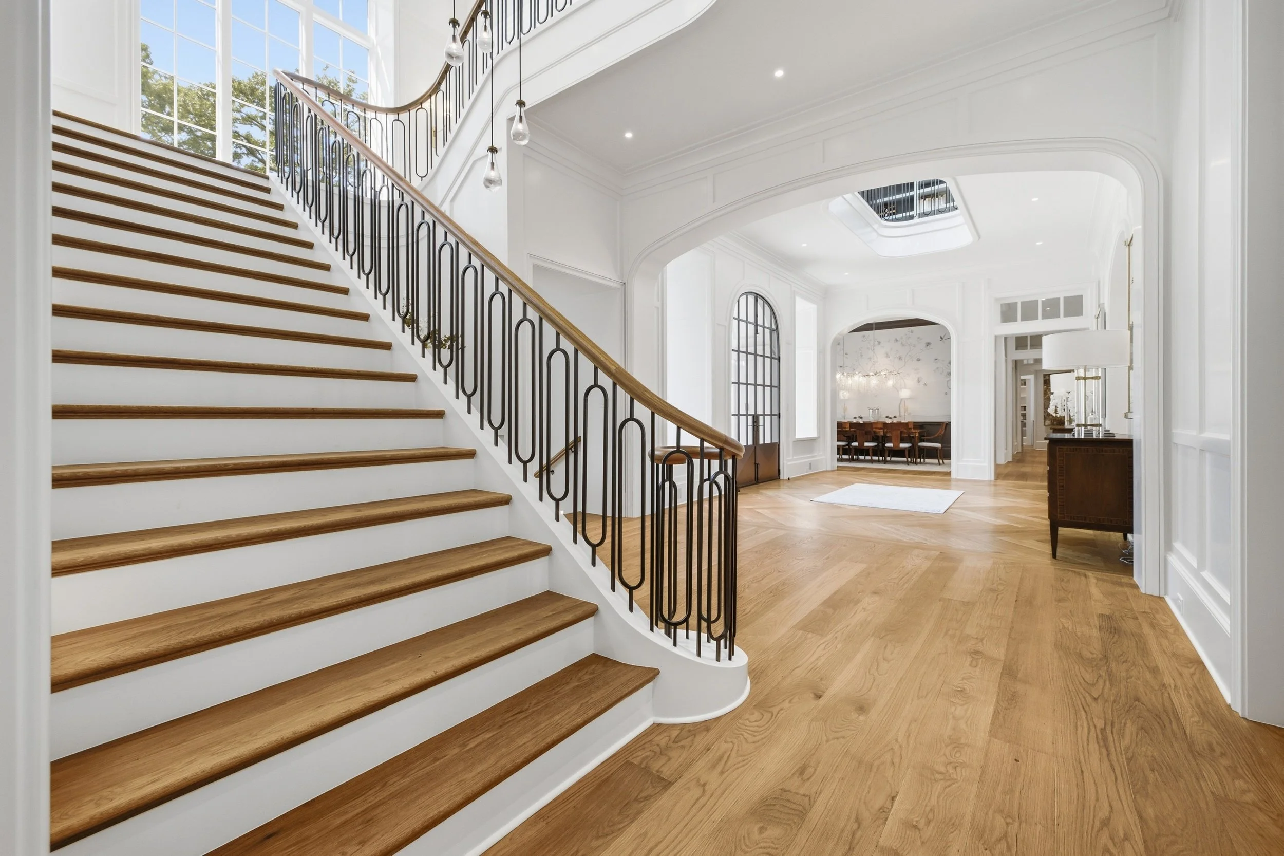 Interior of a spacious, modern home featuring a staircase with wooden steps and black metal railing, large windows letting in natural light, white walls with detailed molding, hardwood floors, and a view into a dining area with a table and chairs.