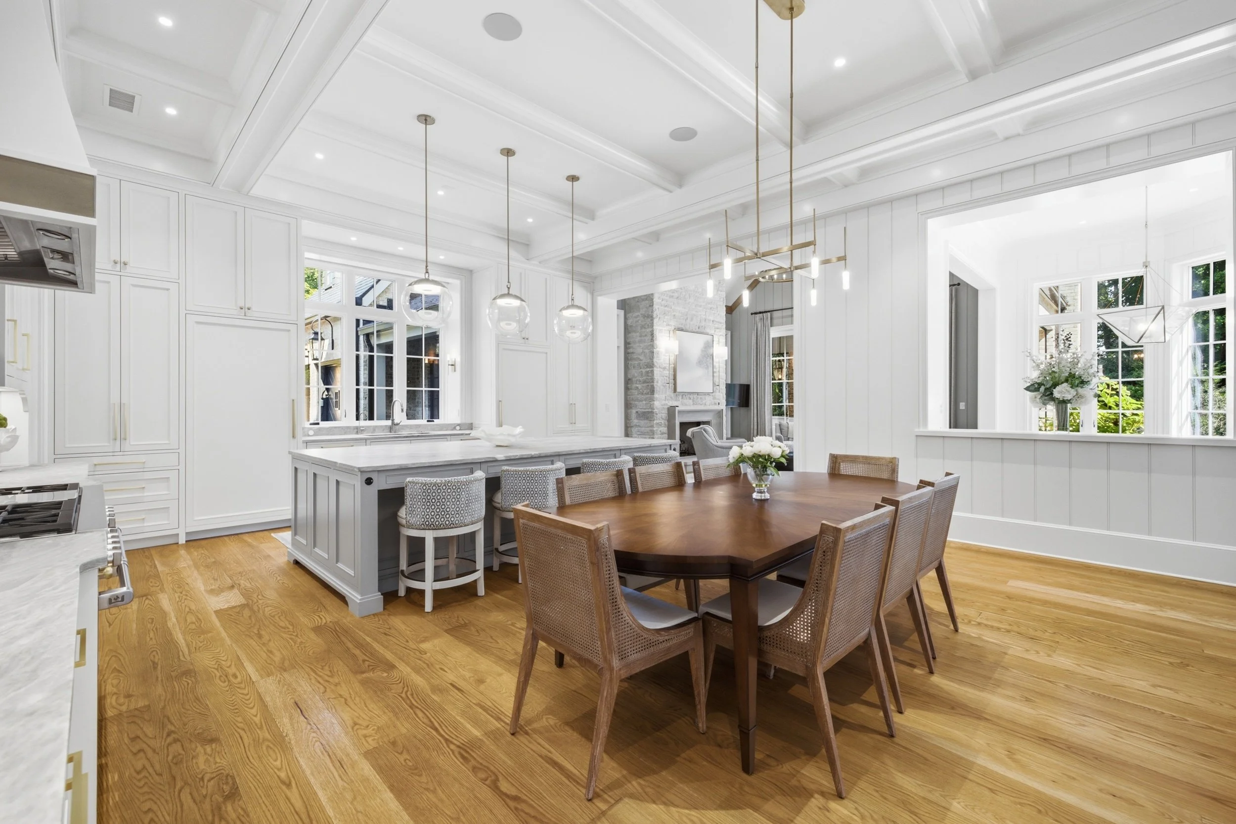 Bright open kitchen and dining area with white cabinetry, wooden floor, and large windows, featuring a kitchen island, hanging pendant lights, and a dining table with chairs, overlooking a living room with a fireplace.