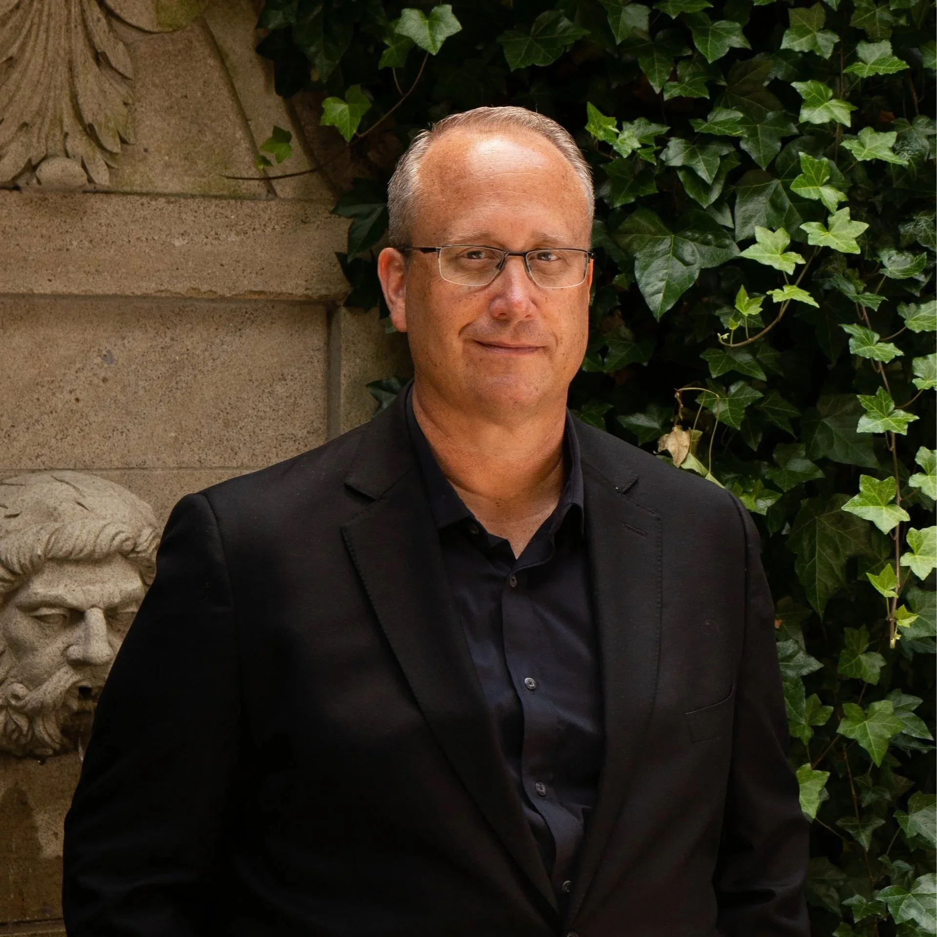A middle-aged man with short, receding hair, wearing glasses, and dressed in a black jacket and shirt, standing outdoors near a stone wall with ivy plants in the background.