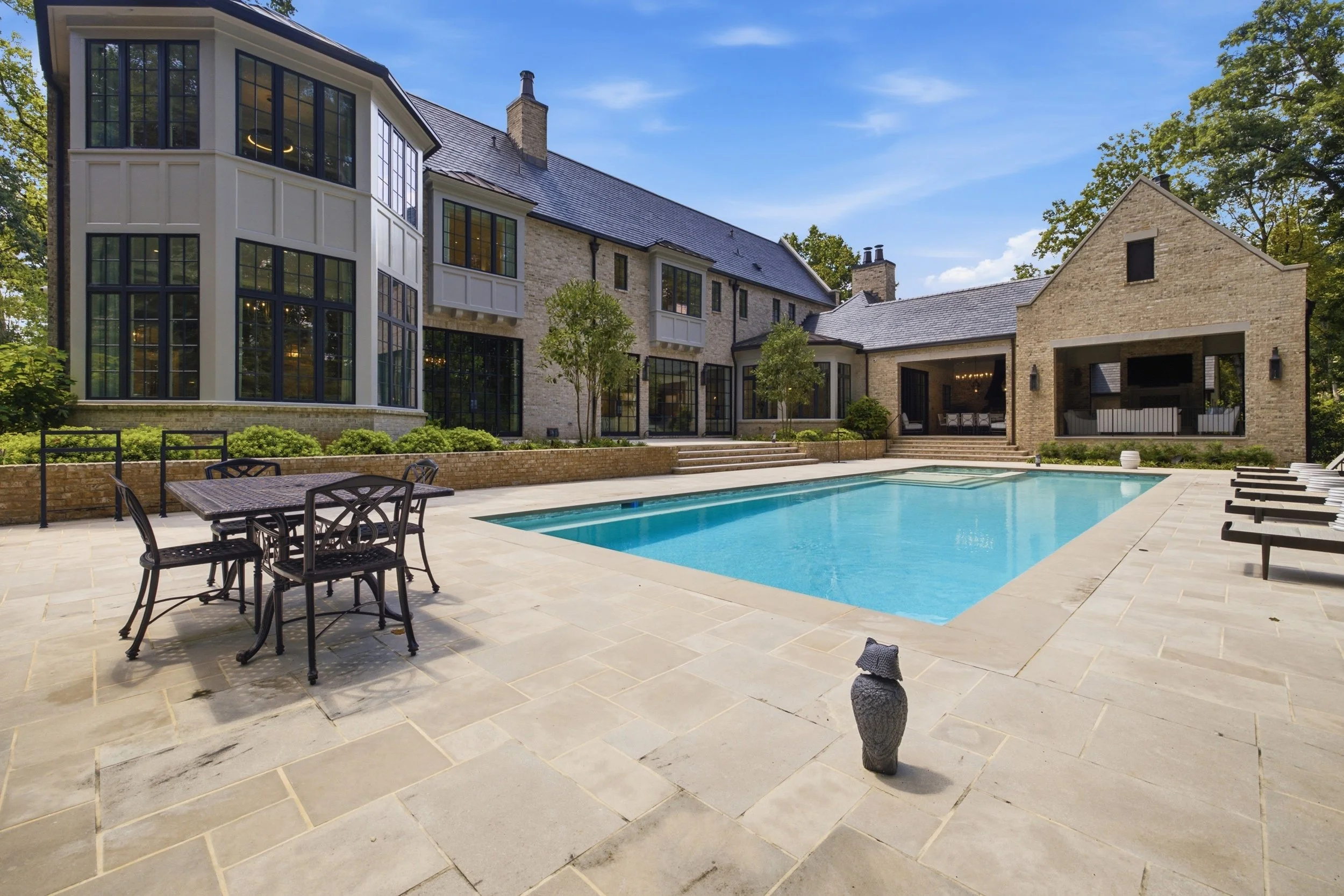 A modern house with large windows and a gray tiled roof, overlooking a backyard with a swimming pool, outdoor furniture, and tall trees under a blue sky.
