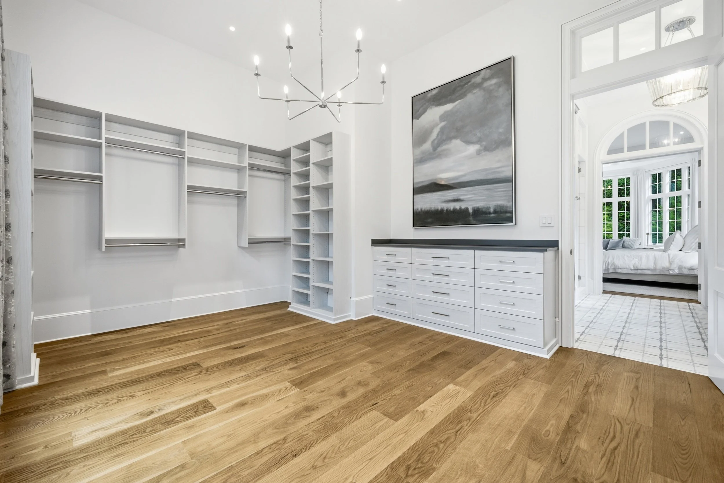 Empty walk-in closet with white shelving, a chandelier, and a bedroom visible through an open door in the background.