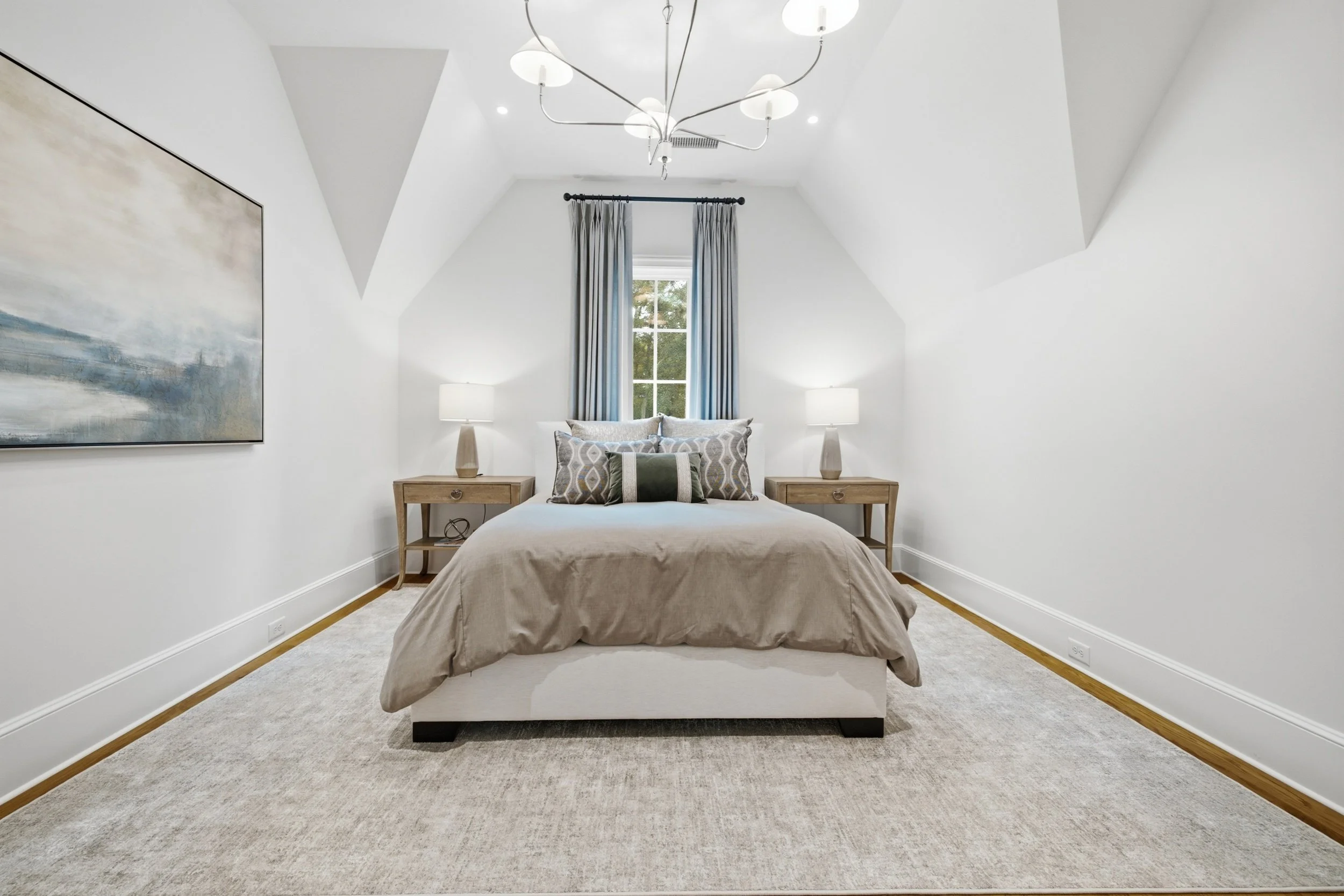 A bedroom with a large window, a bed with beige bedding and multiple pillows, two wooden nightstands with lamps, an abstract painting on the wall, ceiling lights, and gray curtains.