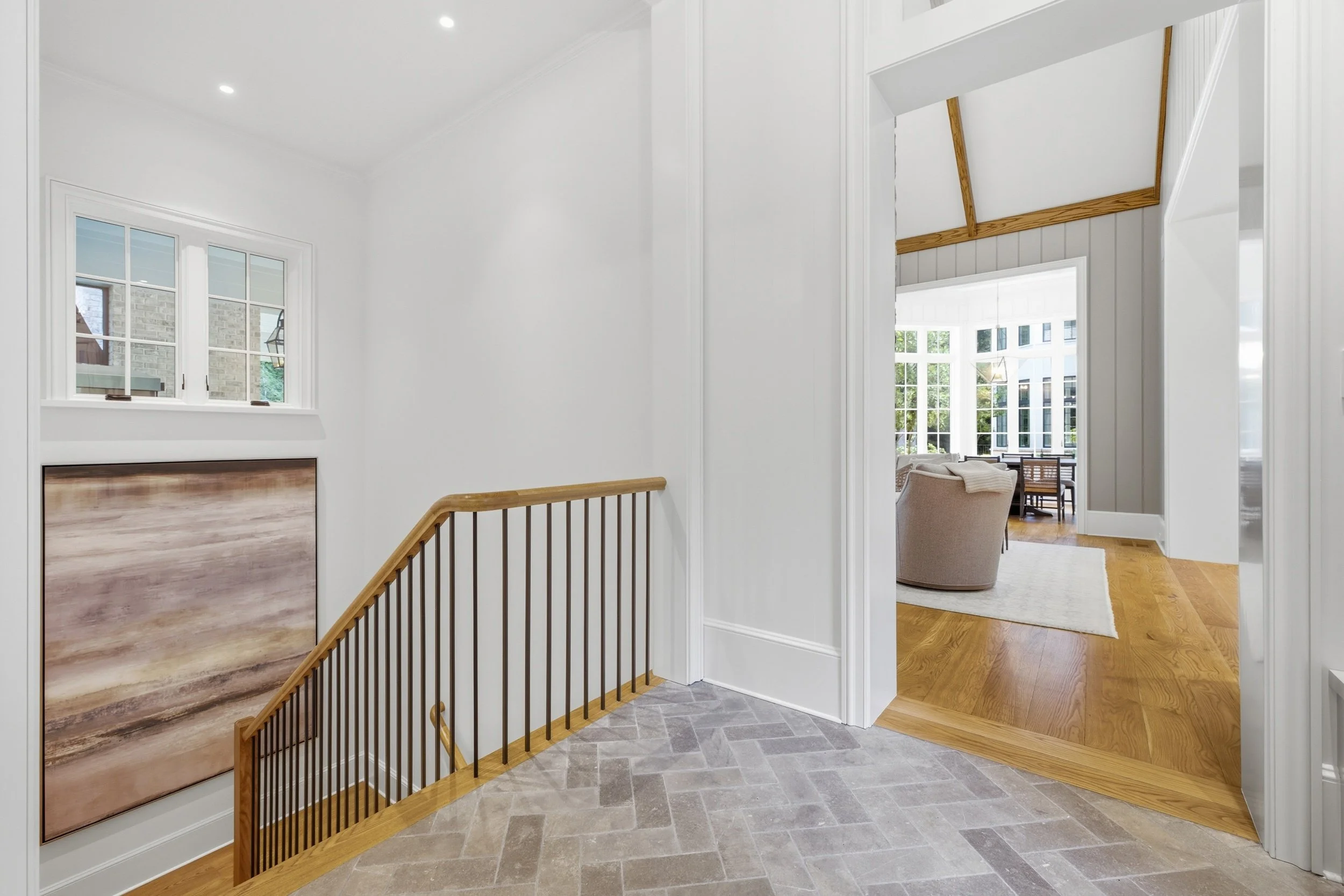 Interior view of a hallway with stairs, white walls, and a glimpse into a bright living room with large windows. The staircase has wooden railings and a wood-paneled storage cabinet under the window. The flooring is a mix of gray tiles and hardwood.