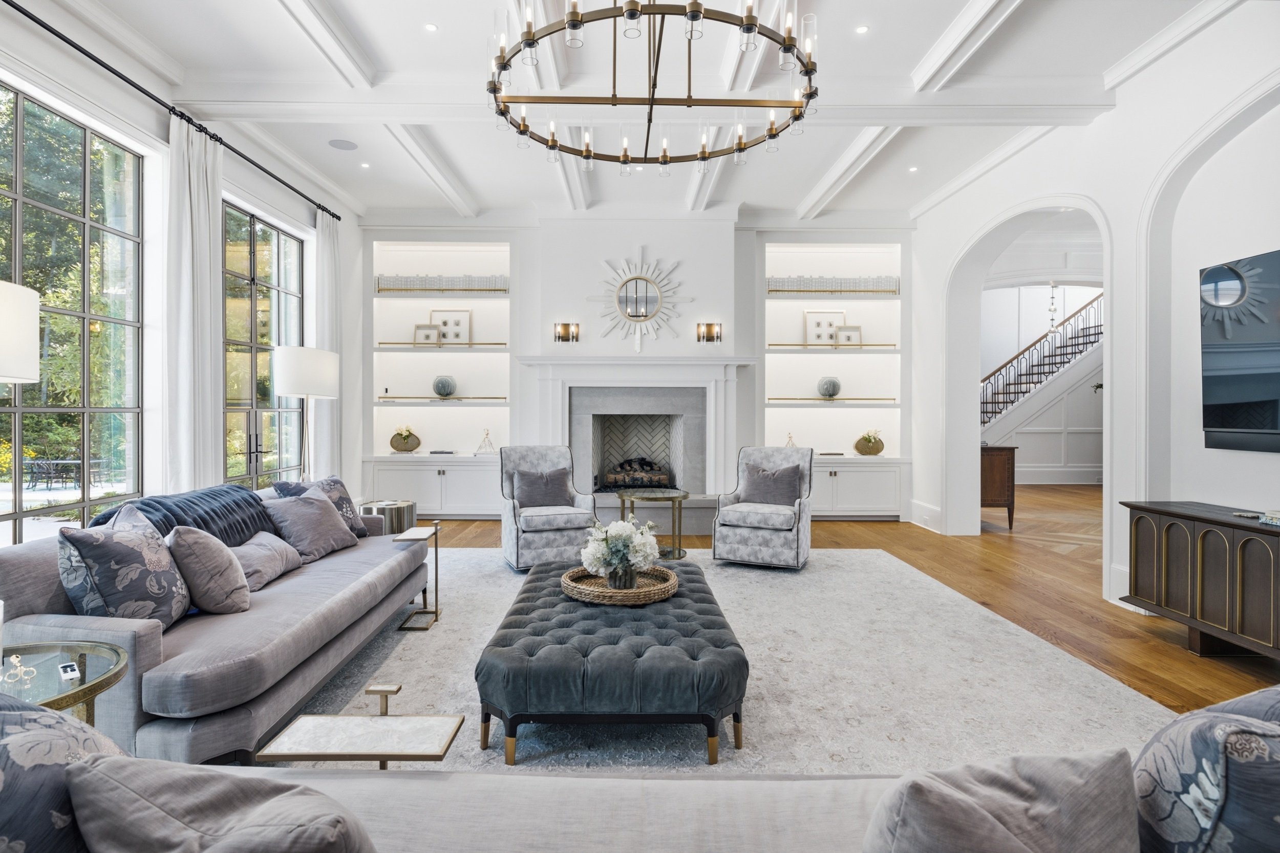 Living room with large windows, white walls, and a fireplace. It features a gray sofa, two gray armchairs, a black tufted ottoman, and a round coffee table with a flower arrangement. There is a gold and glass chandelier hanging from the ceiling.