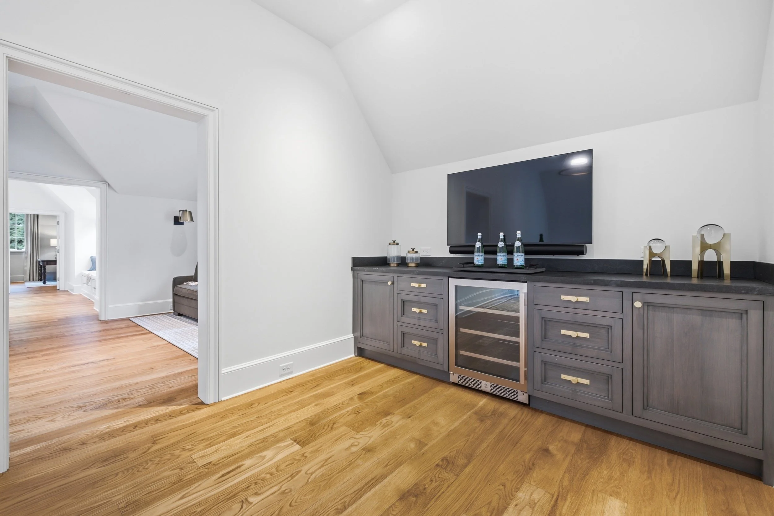 Living room with white walls and a wooden floor, showing a section of a gray cabinet with a built-in wine cooler, glass bottles, decorative items, and a large flat-screen TV wall. There is a doorway leading to another room with a window and a partial