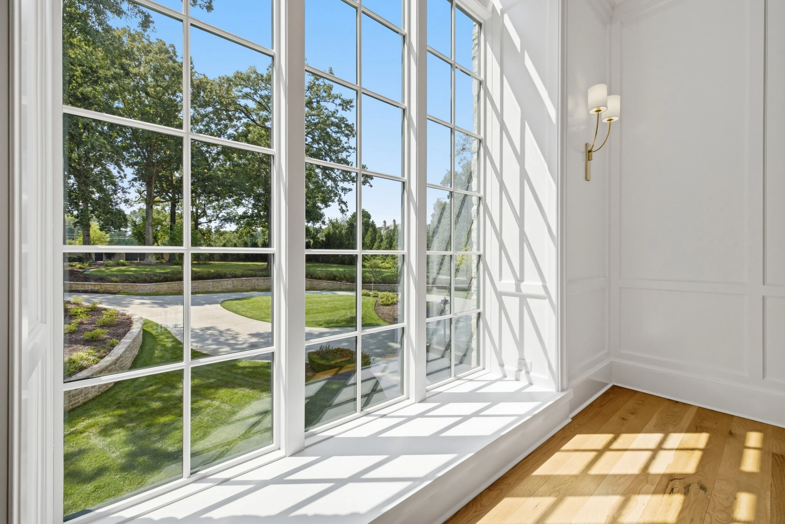 A large window with multiple panes showing a view of a lush green garden and a bright blue sky, inside a white room with wooden flooring and a wall light fixture.