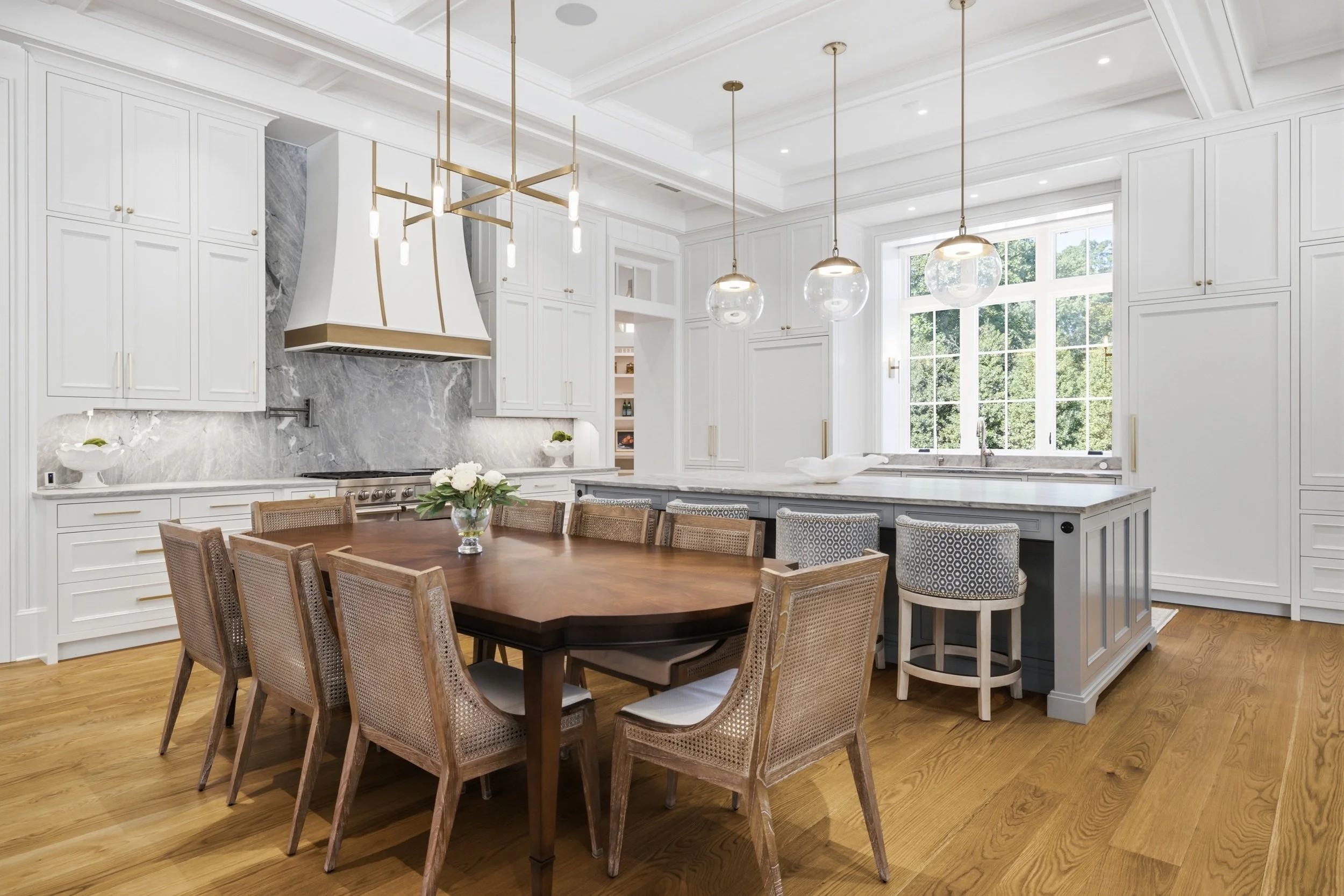 A spacious white kitchen with a large wooden dining table surrounded by twelve chairs, featuring modern pendant and chandelier lighting, with large windows showing greenery outside.