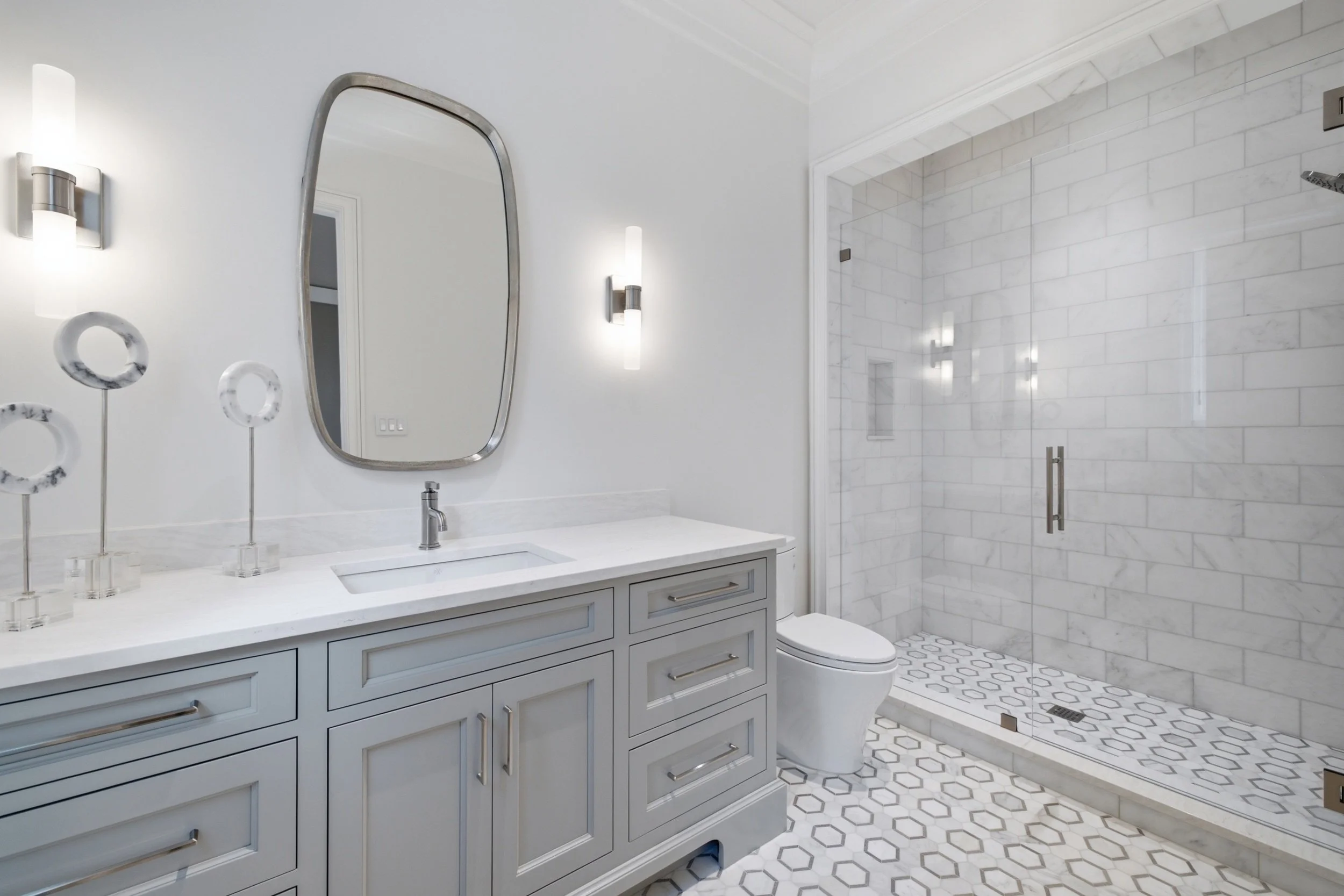 Modern bathroom with white vanity, marble countertop, oval mirror, and shower with glass door and white subway tile walls.
