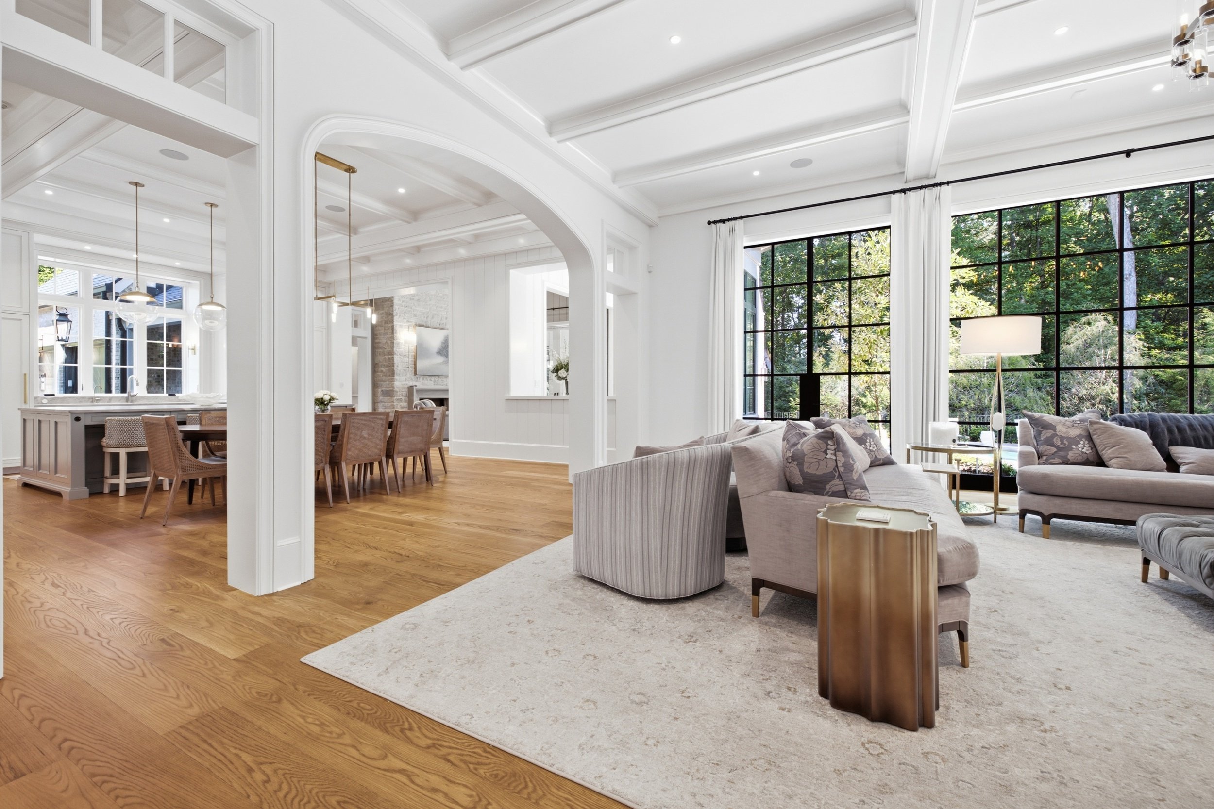 Bright, open living and dining area with large windows, white walls and ceiling, hardwood floors, and modern furniture including a sofa, armchair, and dining table with chairs.