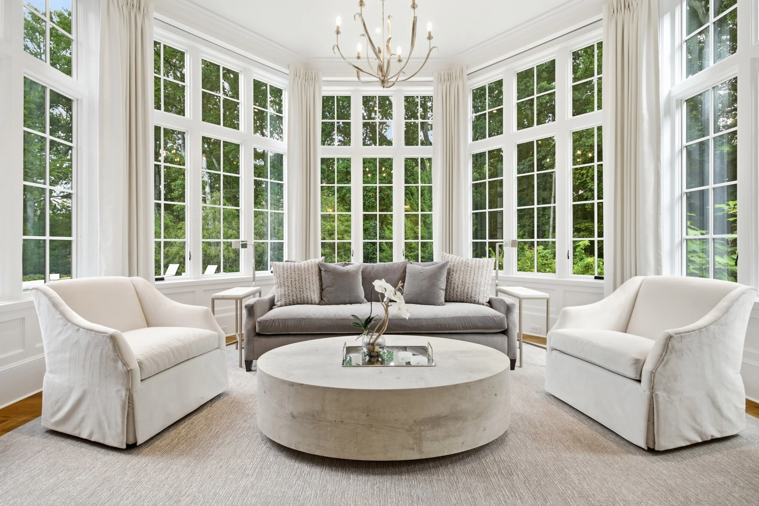 Living room with large bay windows, white curtains, a chandelier, gray sofa with pillows, white armchairs, a round coffee table with a flower arrangement, and greenery visible outside