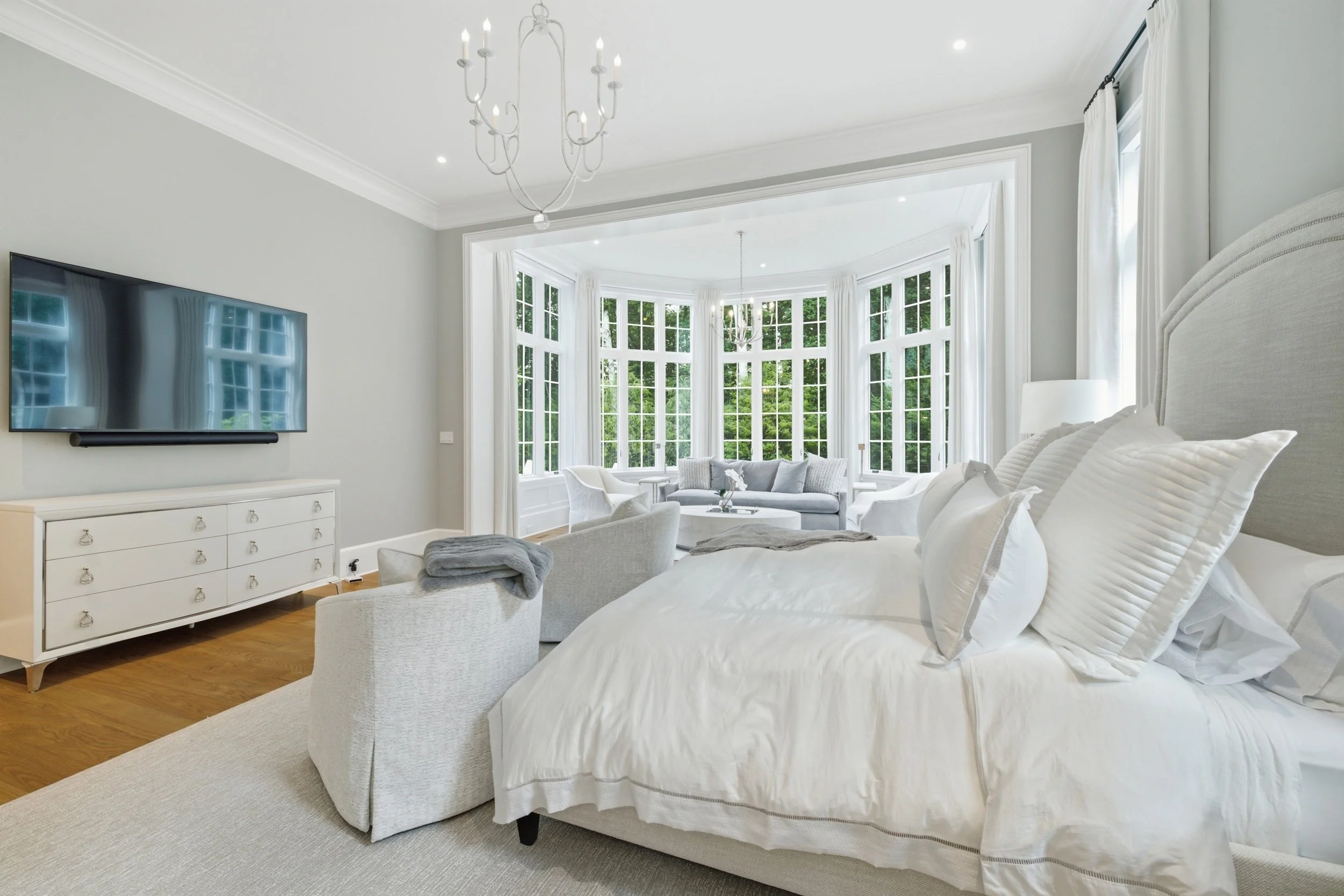 Bright bedroom with large bay windows, white bedding, a dresser with a TV, white curtains, a chandelier, and neutral decor.