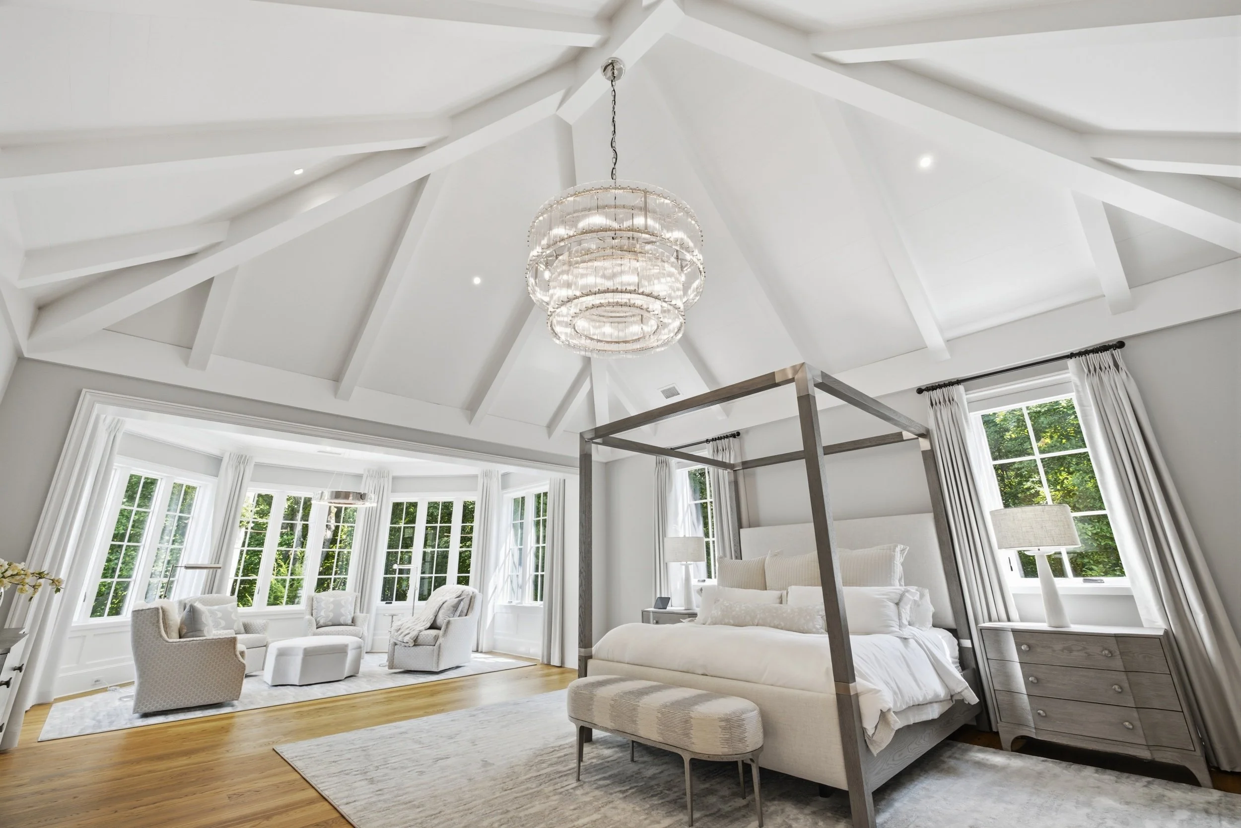 A spacious, bright living room with white vaulted ceiling featuring exposed beams, large windows with white curtains, a crystal chandelier, and white furniture including armchairs, a bed, and side tables.