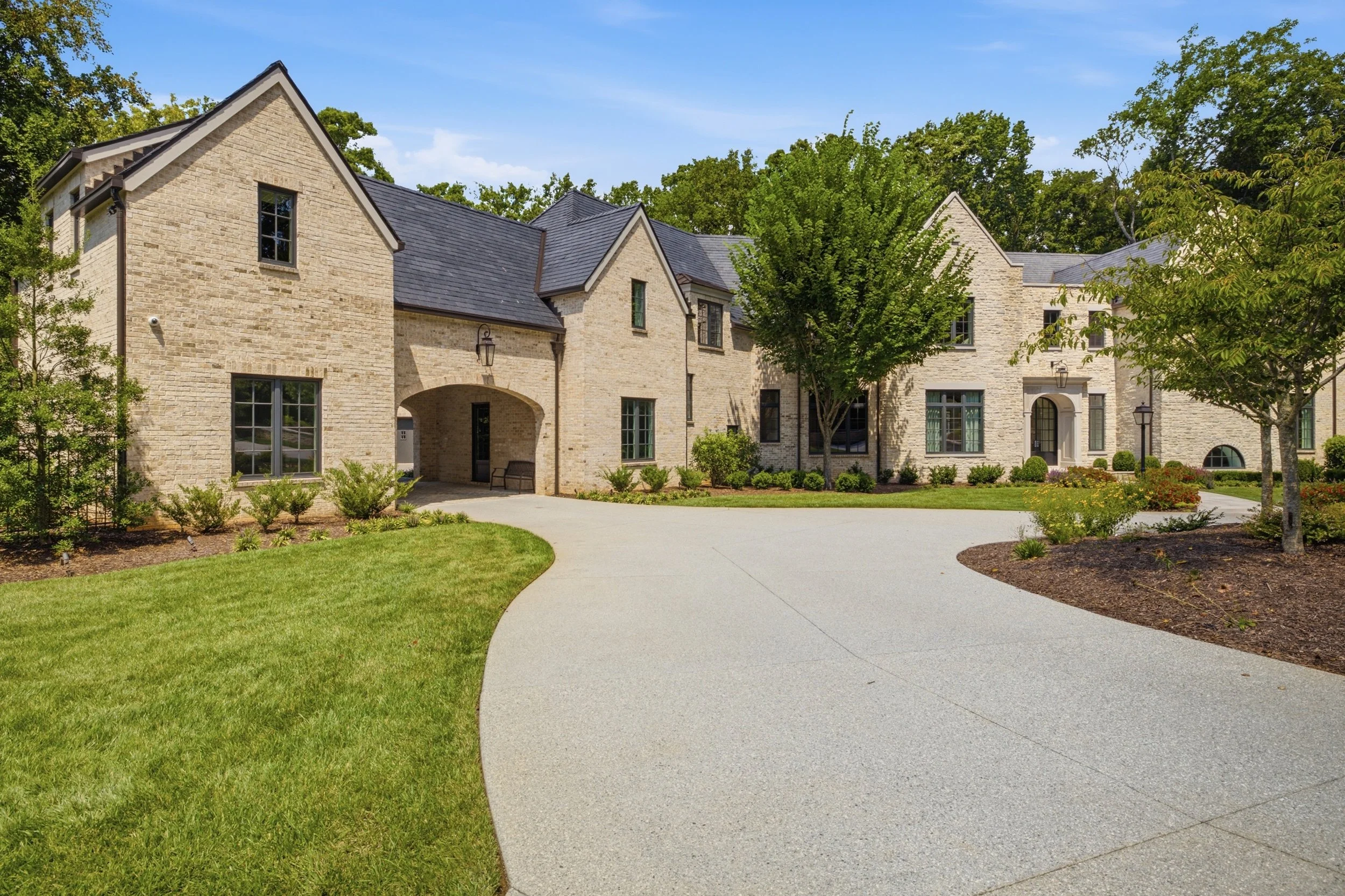 A large brick house with a slate roof, surrounded by green trees and a neatly maintained lawn, with a curved concrete driveway leading to the entrance on a sunny day.