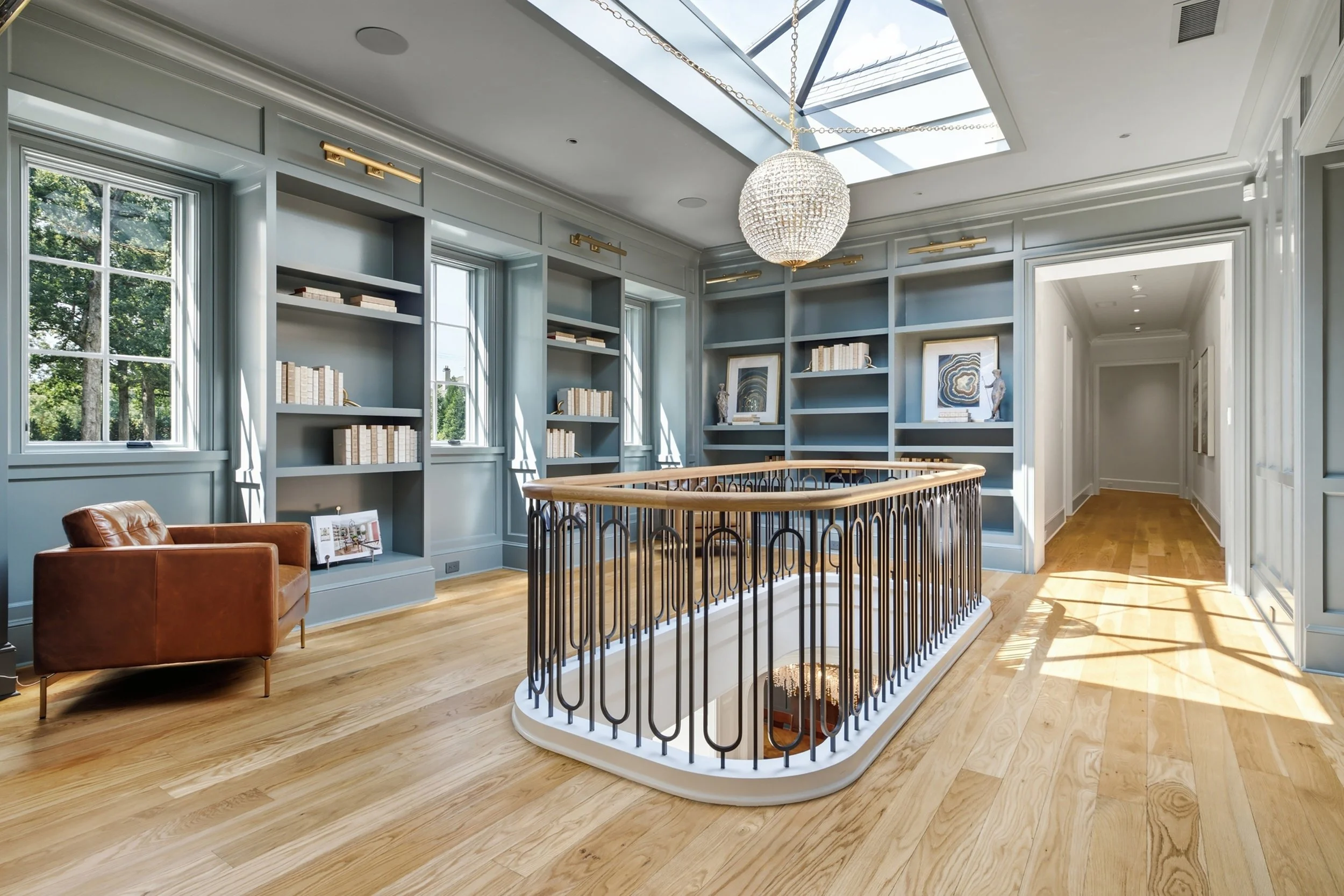 Interior view of a light-filled room with large windows, a chandelier, bookshelves, and a staircase railing, featuring hardwood floors and decorative art.