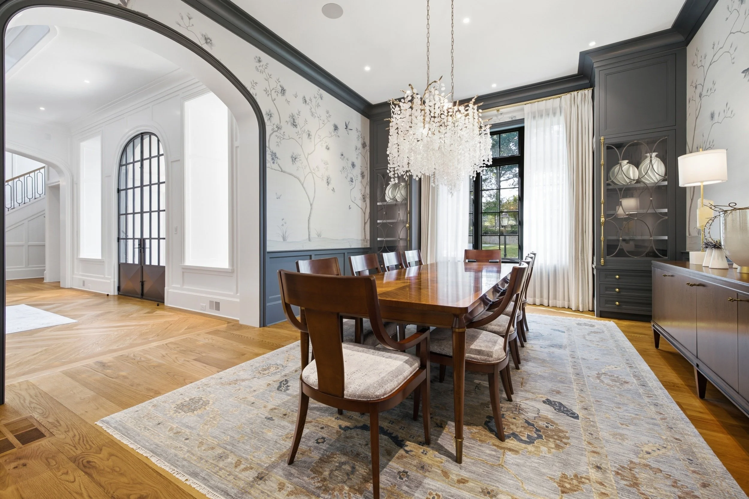 A dining room with a wooden table and eight chairs, a large chandelier, dark gray built-in cabinets, patterned wallpaper, white curtains, and large windows showing a garden outside.