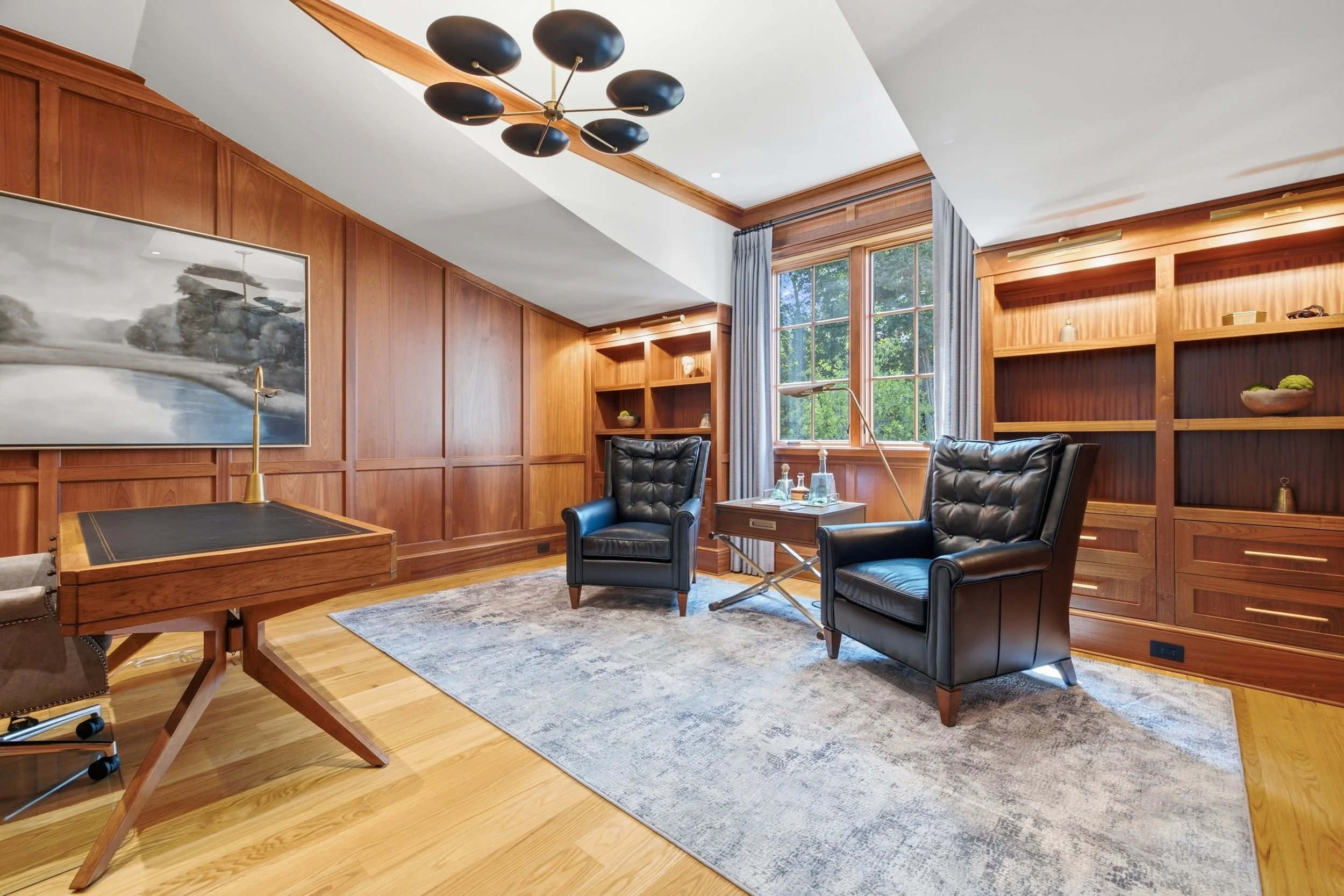 A cozy living room with wood-paneled walls and large windows with blue curtains, furnished with a black leather armchair, a black leather side chair, a wooden desk with brass lamp, and built-in wooden bookshelves with decorative items. A modern chand
