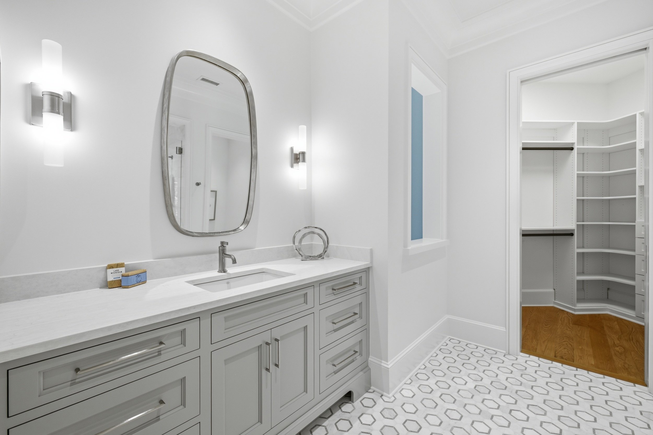 A modern white bathroom with a vanity, marble countertop, and elongated mirror. There are wall-mounted light fixtures, and an open walk-in closet with white shelving and wood flooring.