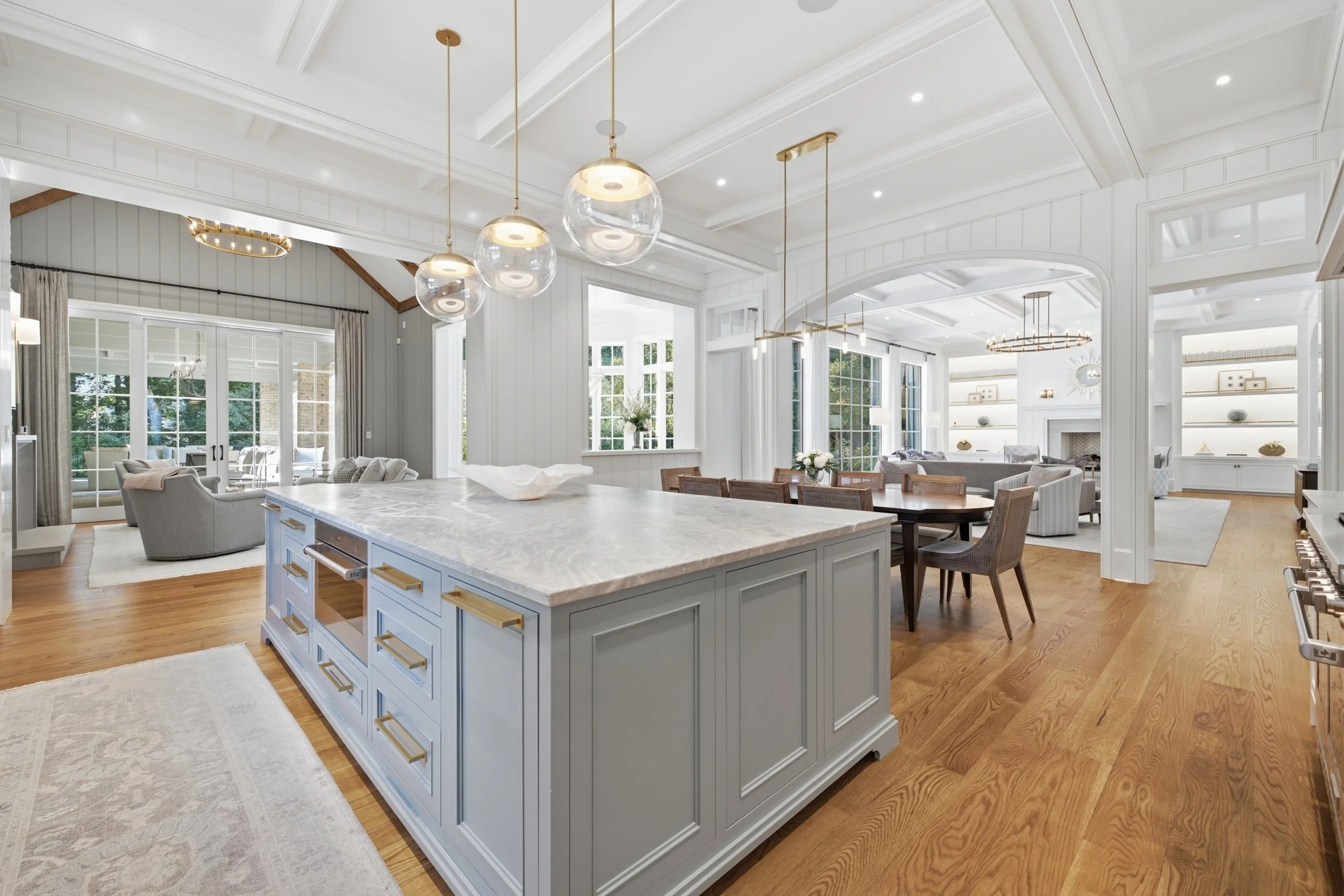 Bright and spacious living area with large windows, white walls, and detailed ceiling design, featuring a kitchen island with a marble top, pendant lighting, and seating area with sofas and chairs.