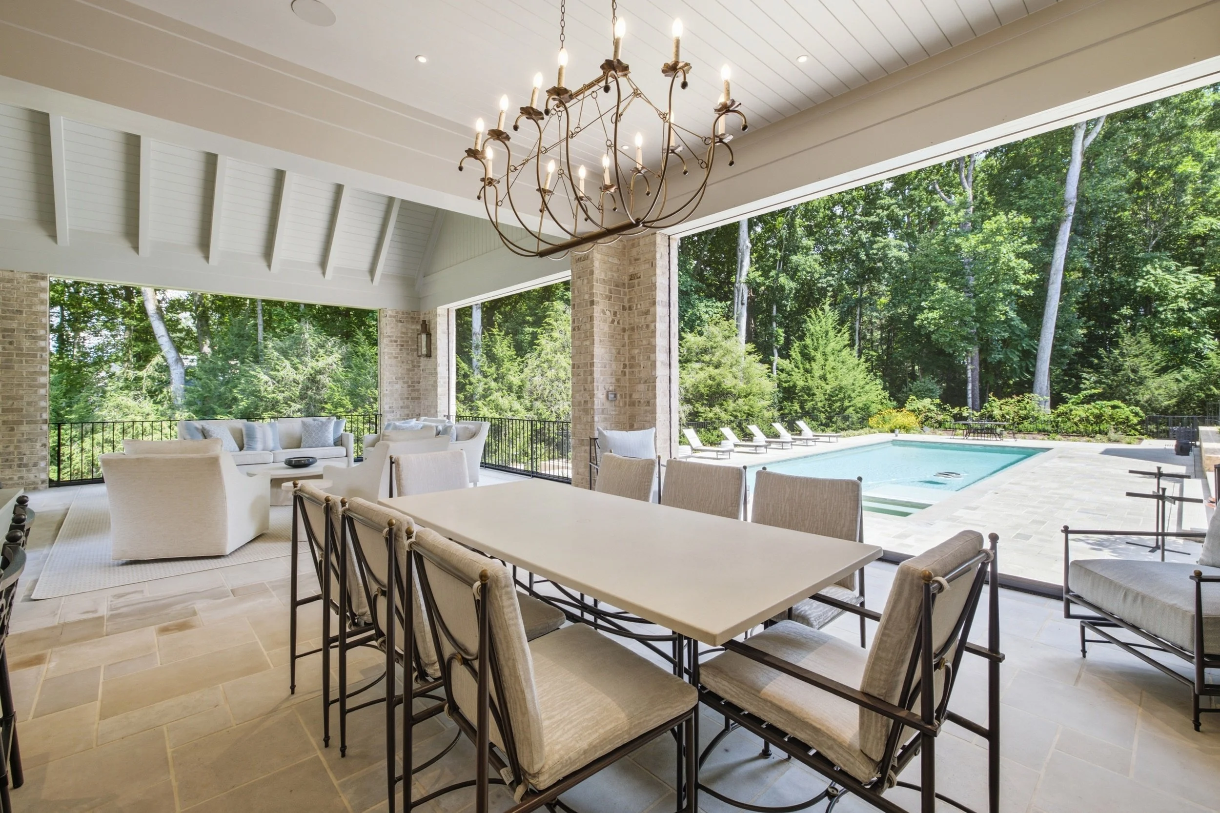 Covered outdoor patio with a large dining table, multiple chairs, a chandelier, a seating area with sofas and cushions, a swimming pool, and lounge chairs, surrounded by green trees.