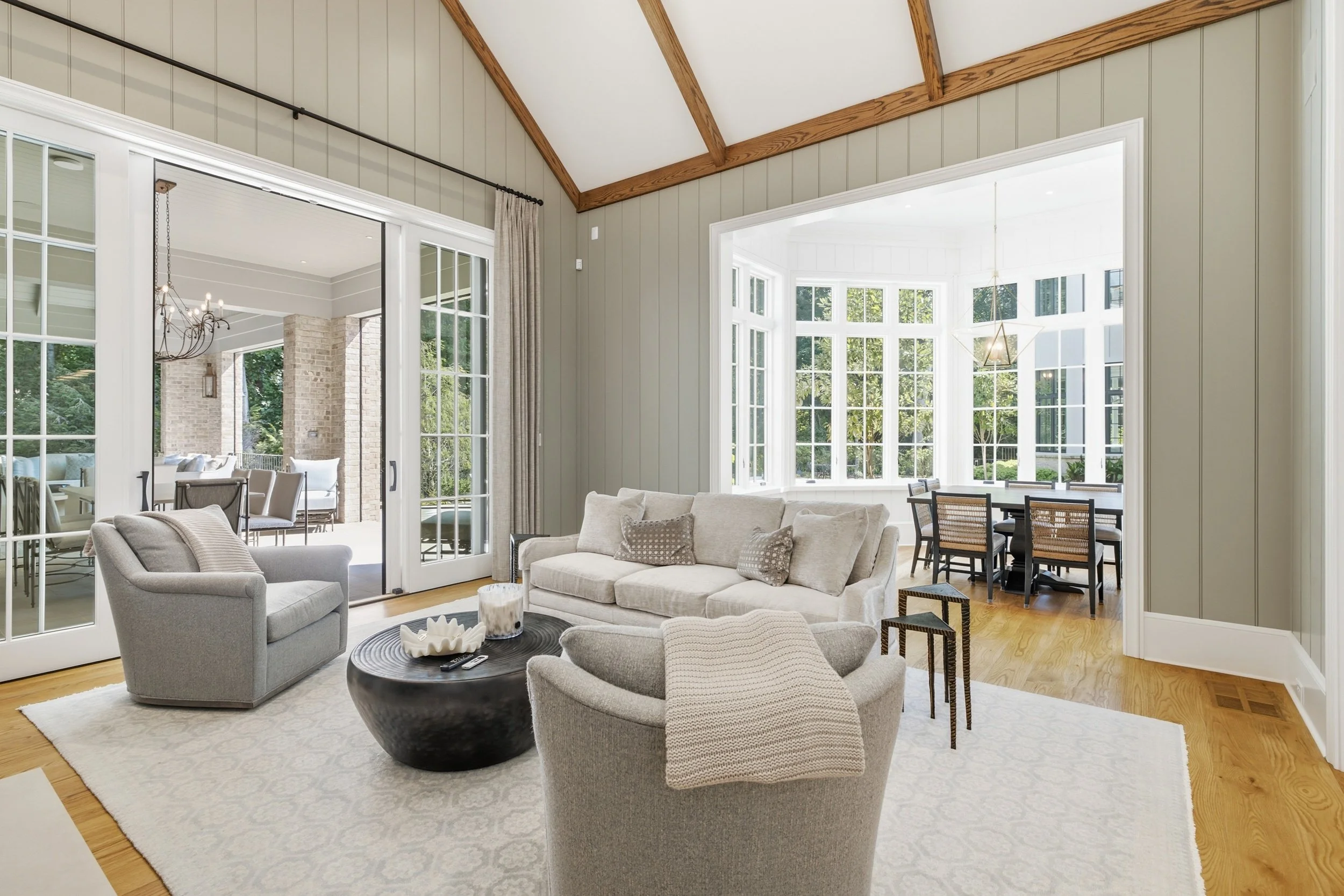 Living room with beige sofa, armchair, black coffee table, and round dining table with chairs, large windows, and a sliding glass door leading to an outdoor patio with seating.